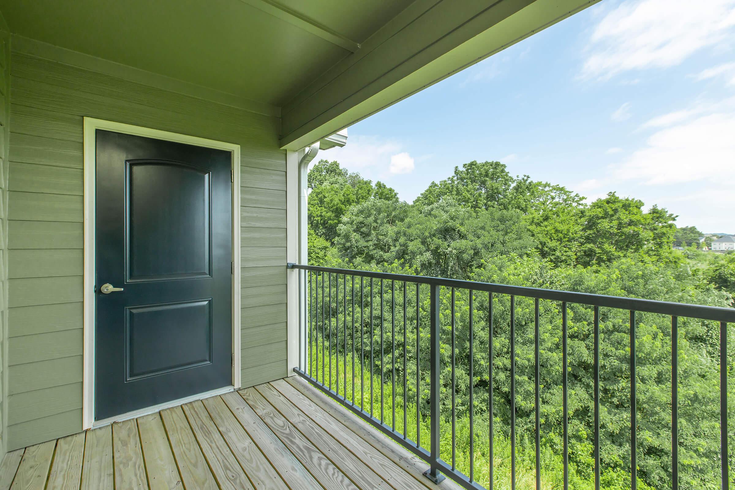 A view from a balcony with a dark door leading indoors. The wooden floor is visible, and lush green trees are seen in the background under a partly cloudy blue sky. The balcony railing frames the view, creating an inviting outdoor space.