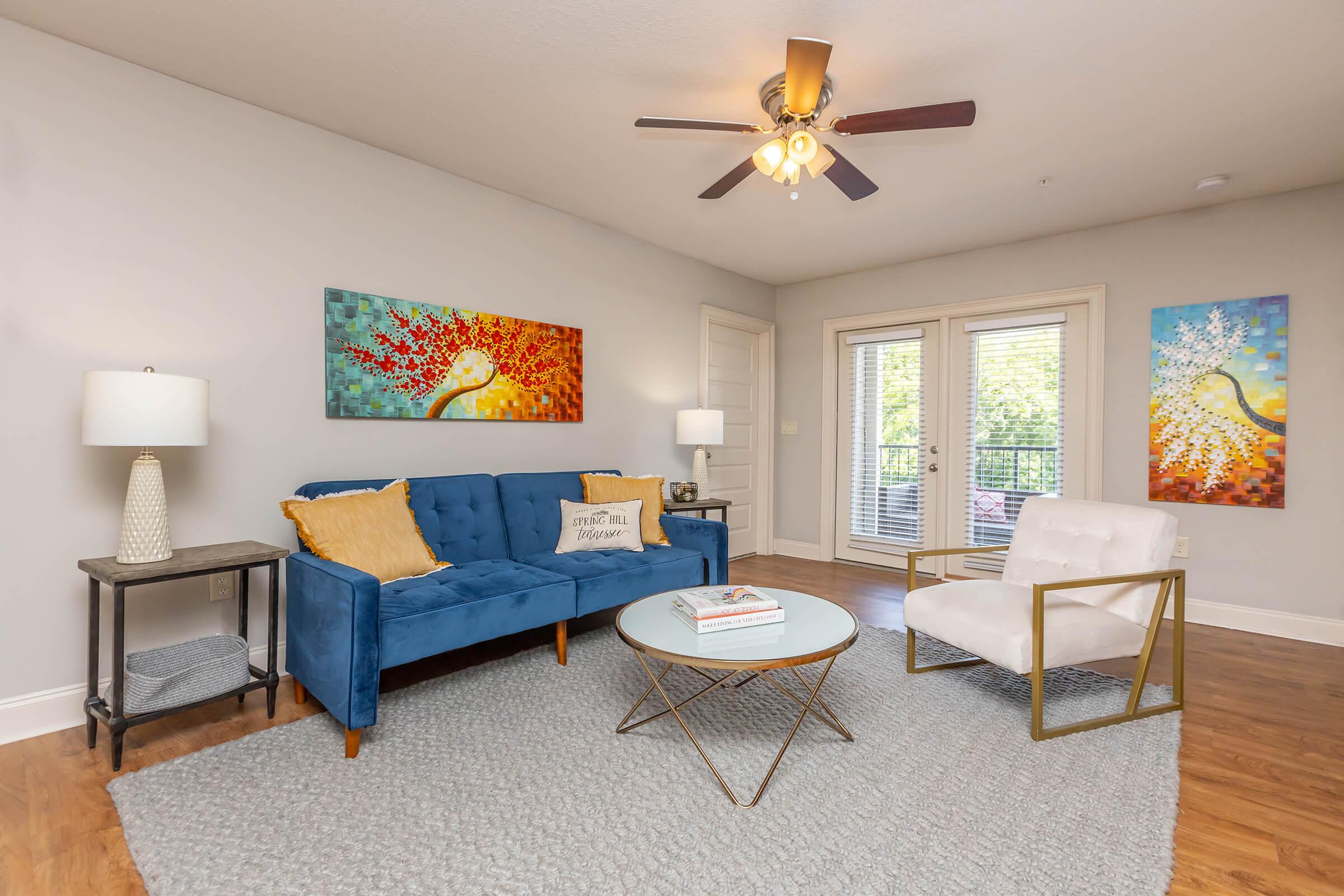 A cozy living room featuring a blue couch with yellow and white pillows, a round coffee table with a book, and two colorful abstract paintings on the walls. There are two lamps on either side of the couch, and large windows allow natural light to enter the space.