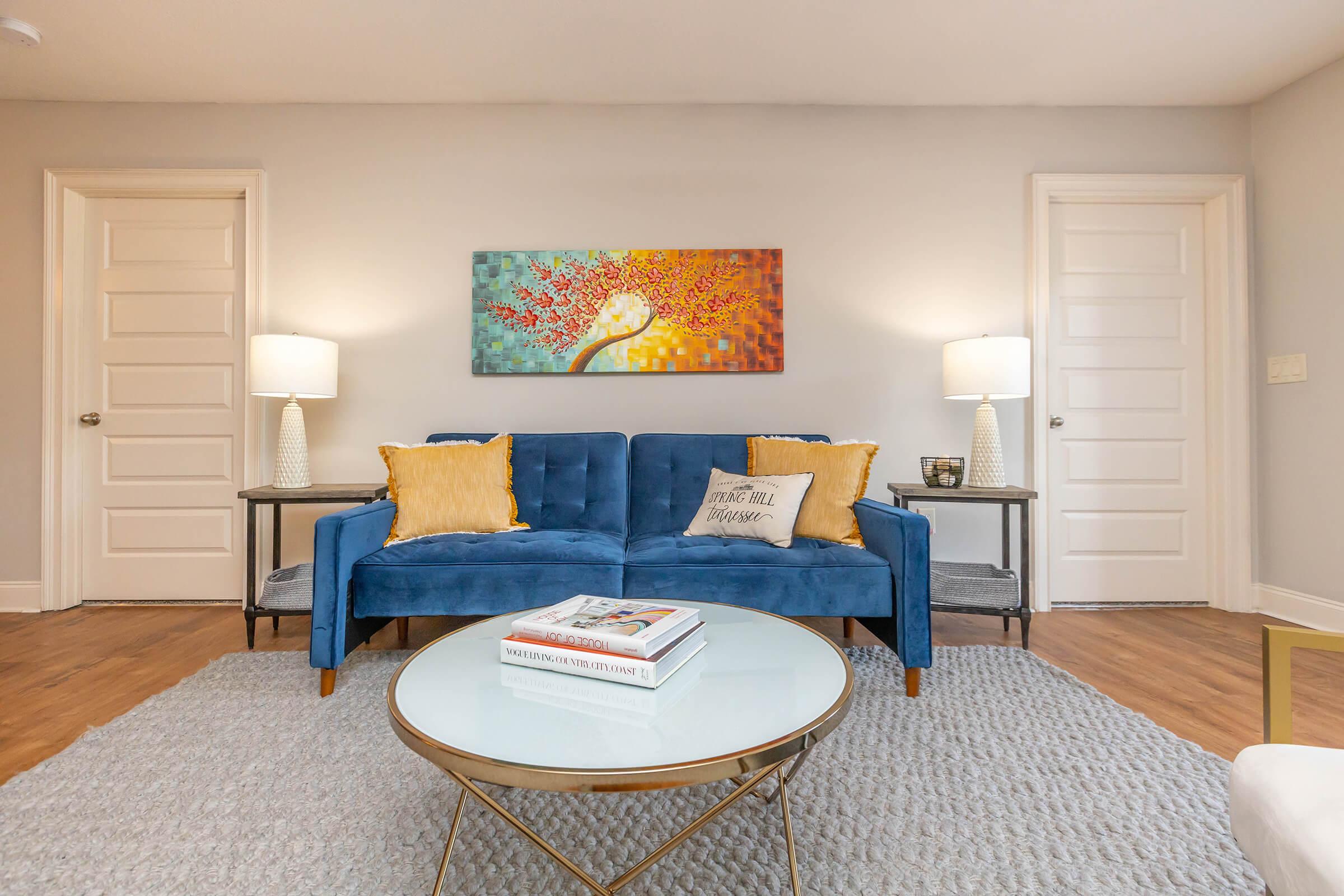 A cozy living room decorated with a blue sofa adorned with yellow pillows. There is a round glass coffee table in front, featuring books and a decorative item. The walls are light gray, and an abstract painting with warm colors hangs above the sofa. Two stylish lamps provide soft lighting.