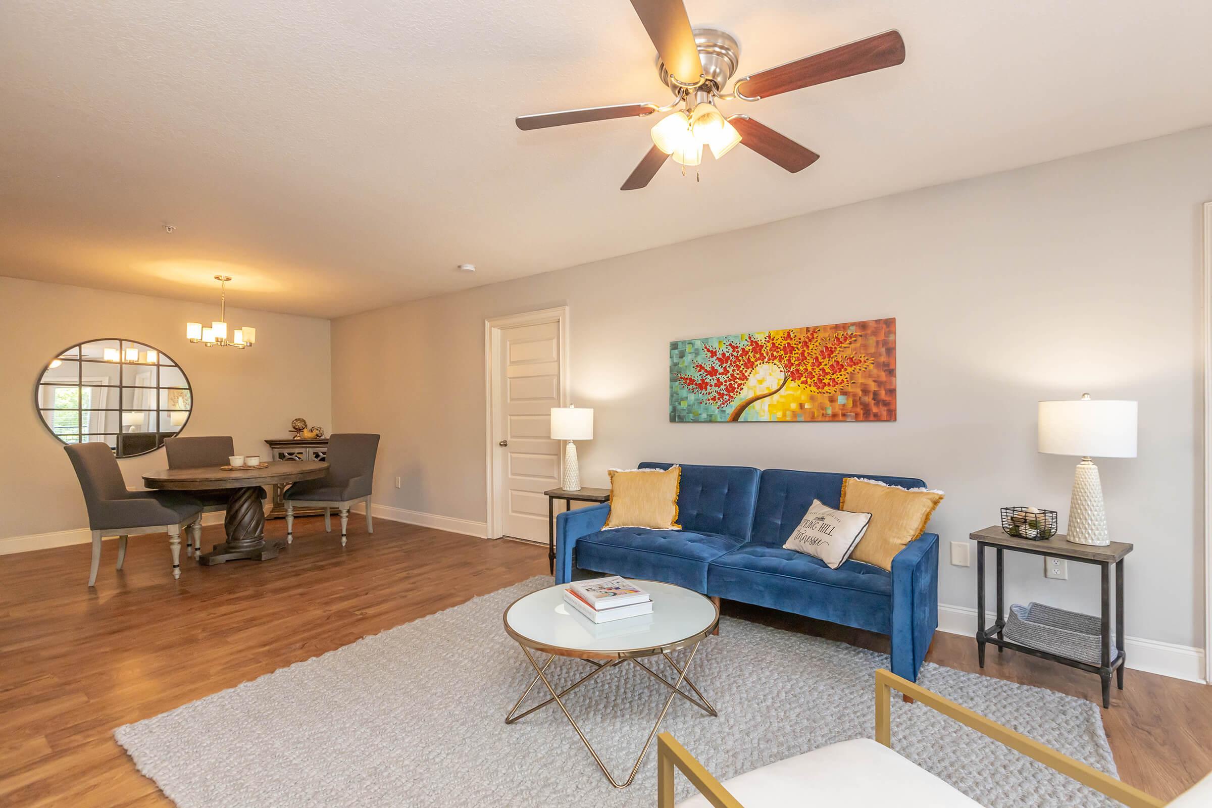 Cozy living space featuring a blue velvet sofa with decorative cushions, a round coffee table, and a textured area rug. A ceiling fan hangs above, and there’s a modern chair in the foreground. In the background, a dining area with a round table and stylish chairs is visible, along with a piece of colorful artwork on the wall.