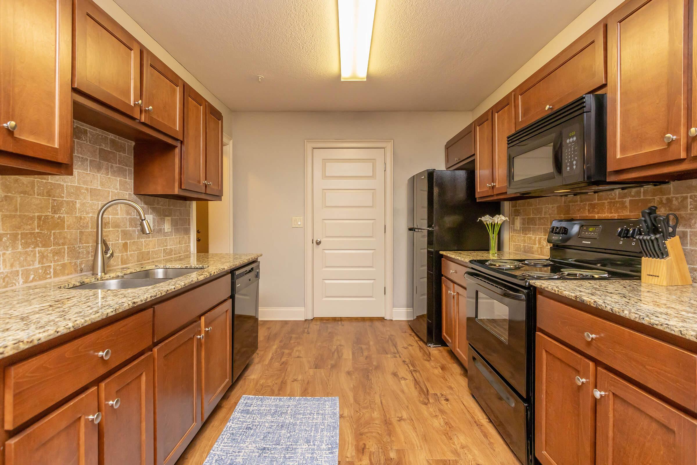 A modern kitchen featuring wooden cabinets, granite countertops, and stainless steel appliances. The space includes a sink, stove, microwave, and refrigerator, with a light-colored wall and a blue area rug on the wooden floor. A door leads to another room, adding to the kitchen's functionality.