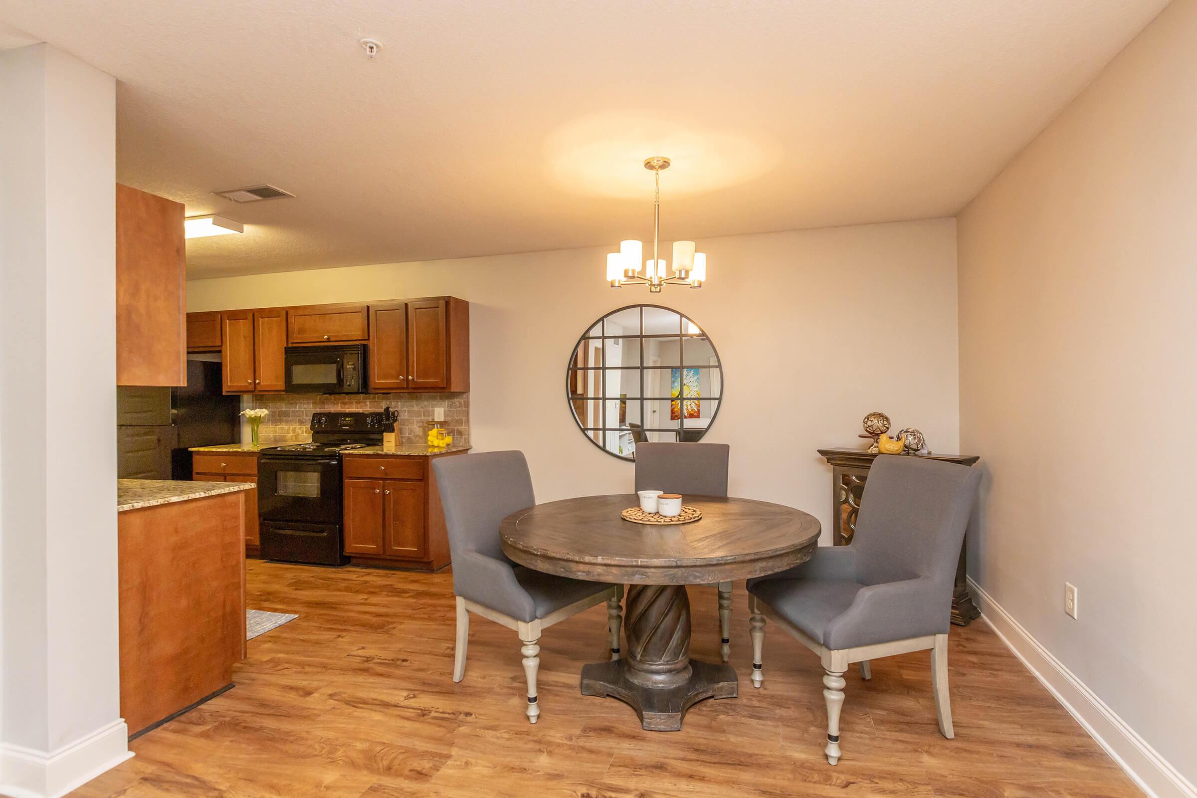 A modern dining area featuring a round wooden table with a decorative centerpiece, surrounded by two gray upholstered chairs. A stylish chandelier hangs above the table. In the background, there's a kitchen with dark wood cabinets and black appliances, and a circular mirror reflecting the space.