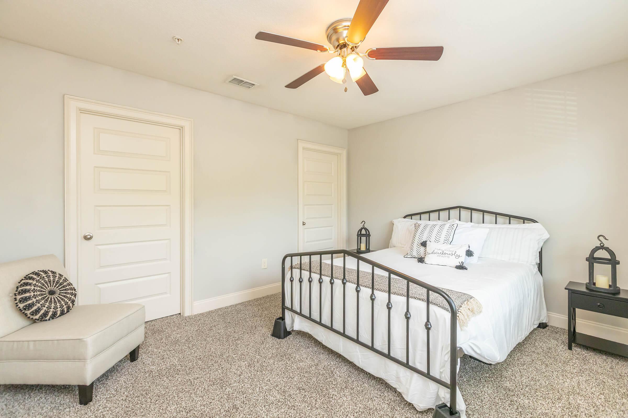 A bright and airy bedroom featuring a metal bed frame with white bedding, a decorative pillow, two doors, and a small black nightstand with a lantern. A light-colored armchair is positioned next to the bed, and the carpeted floor adds warmth to the space. The ceiling fan provides a cozy ambiance.