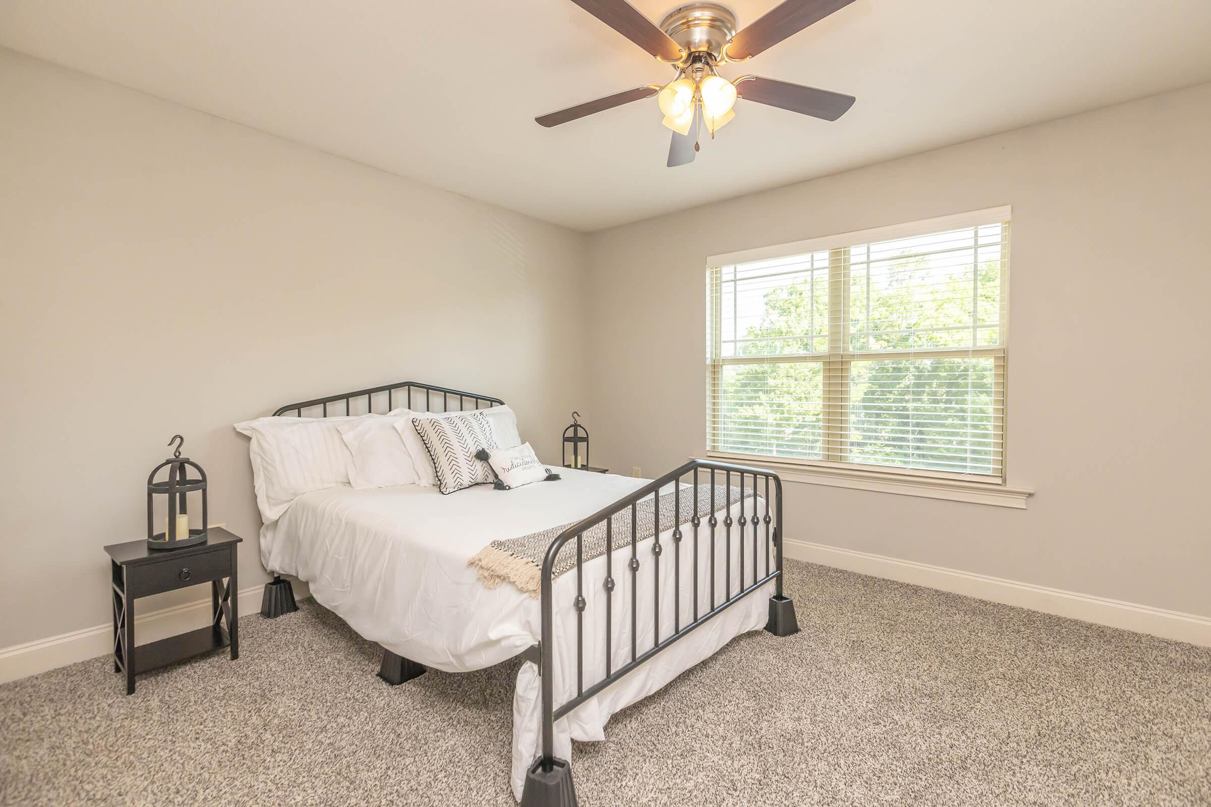 A cozy bedroom featuring a metal bed frame with white bedding, two bedside lanterns on each side, and a ceiling fan. Natural light streams through a large window with blinds, illuminating the soft gray walls and plush carpet flooring, creating a serene and inviting atmosphere.