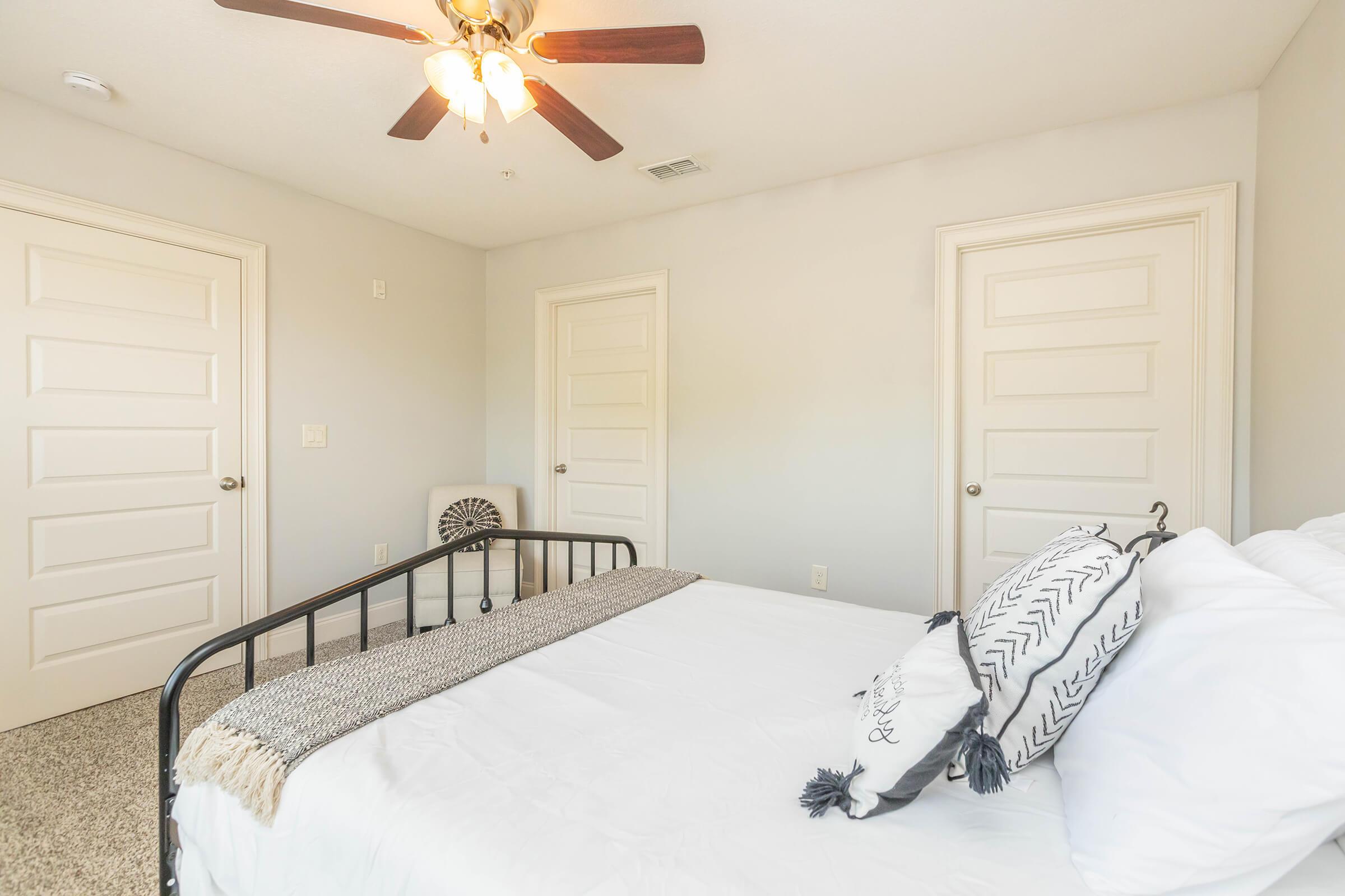 A well-lit bedroom featuring a black metal bed frame with white bedding. There are decorative pillows on the bed and a woven throw blanket at the foot. The room has light gray walls, short carpet, and two closed doors in the background. A ceiling fan is overhead, providing a cozy atmosphere.