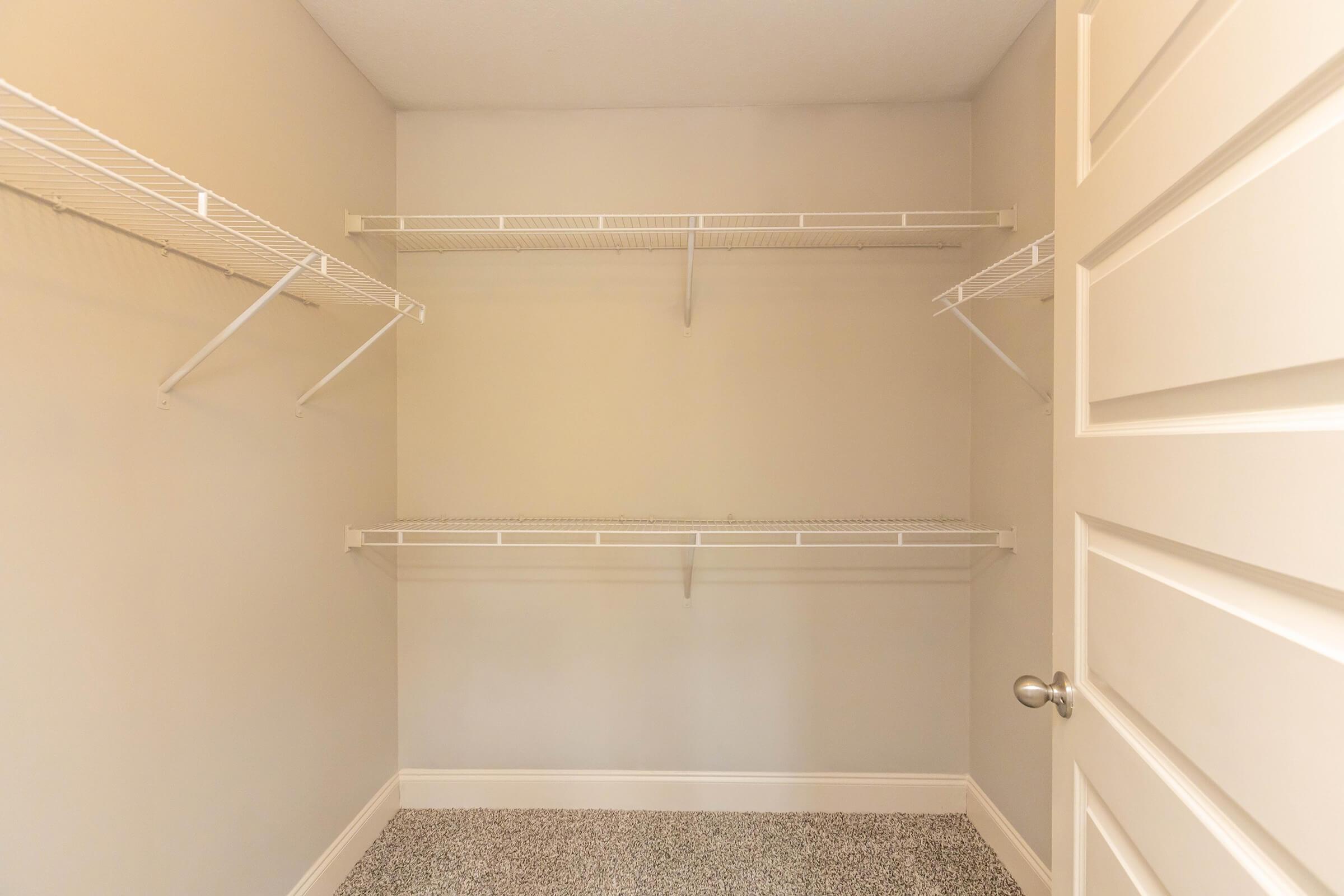 A vacant walk-in closet featuring two wire shelving units mounted on light-colored walls. The floor is covered with a speckled gray material, and a closed white door is visible on the right side. The overall space is well-lit and organized, ready for storage.