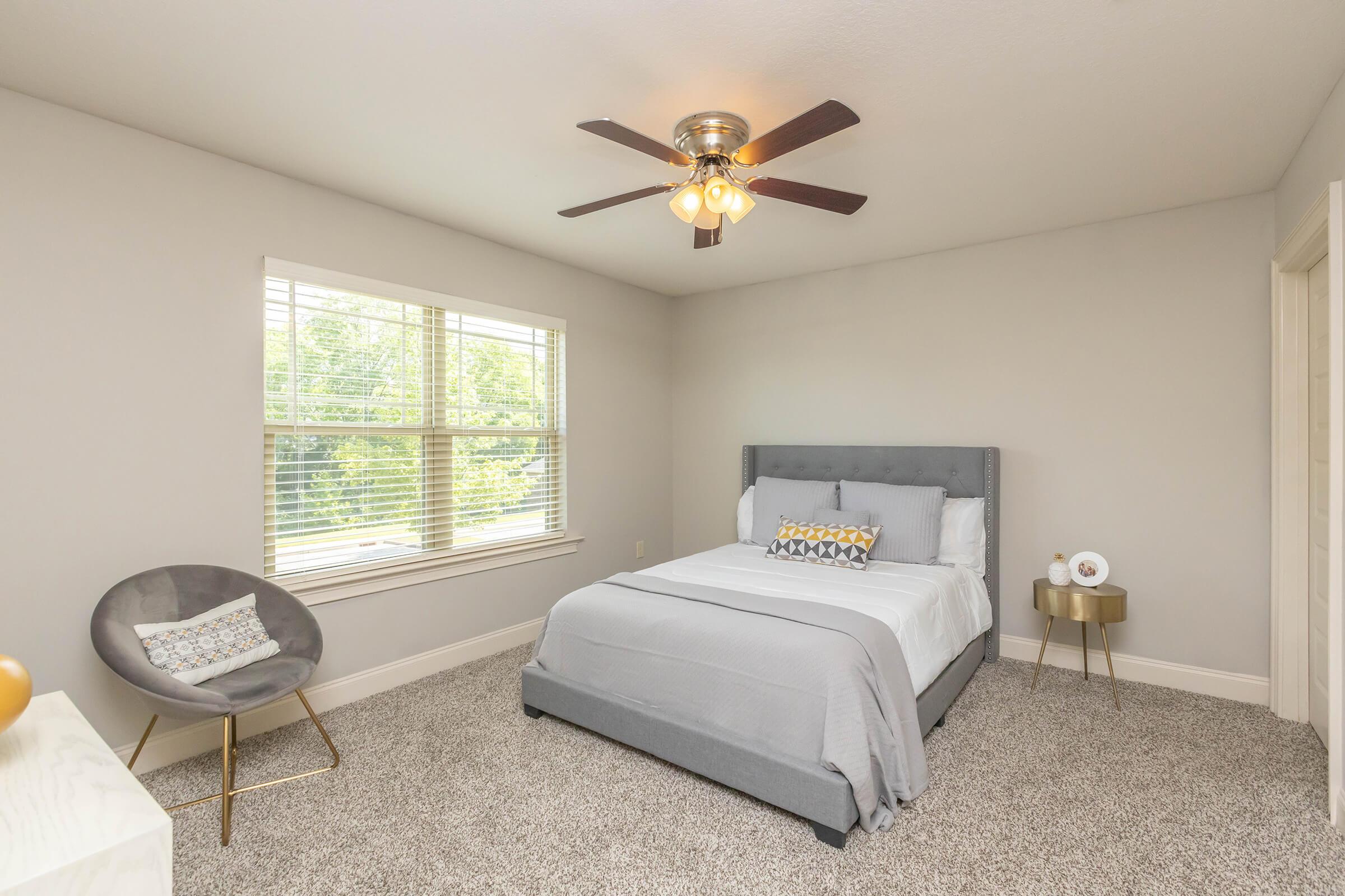 A modern bedroom featuring a gray upholstered bed with a light gray blanket and decorative pillows. There’s a comfortable gray chair beside a small wooden side table, and a ceiling fan above. Natural light streams through a window, illuminating the soft beige walls and plush carpeted floor.