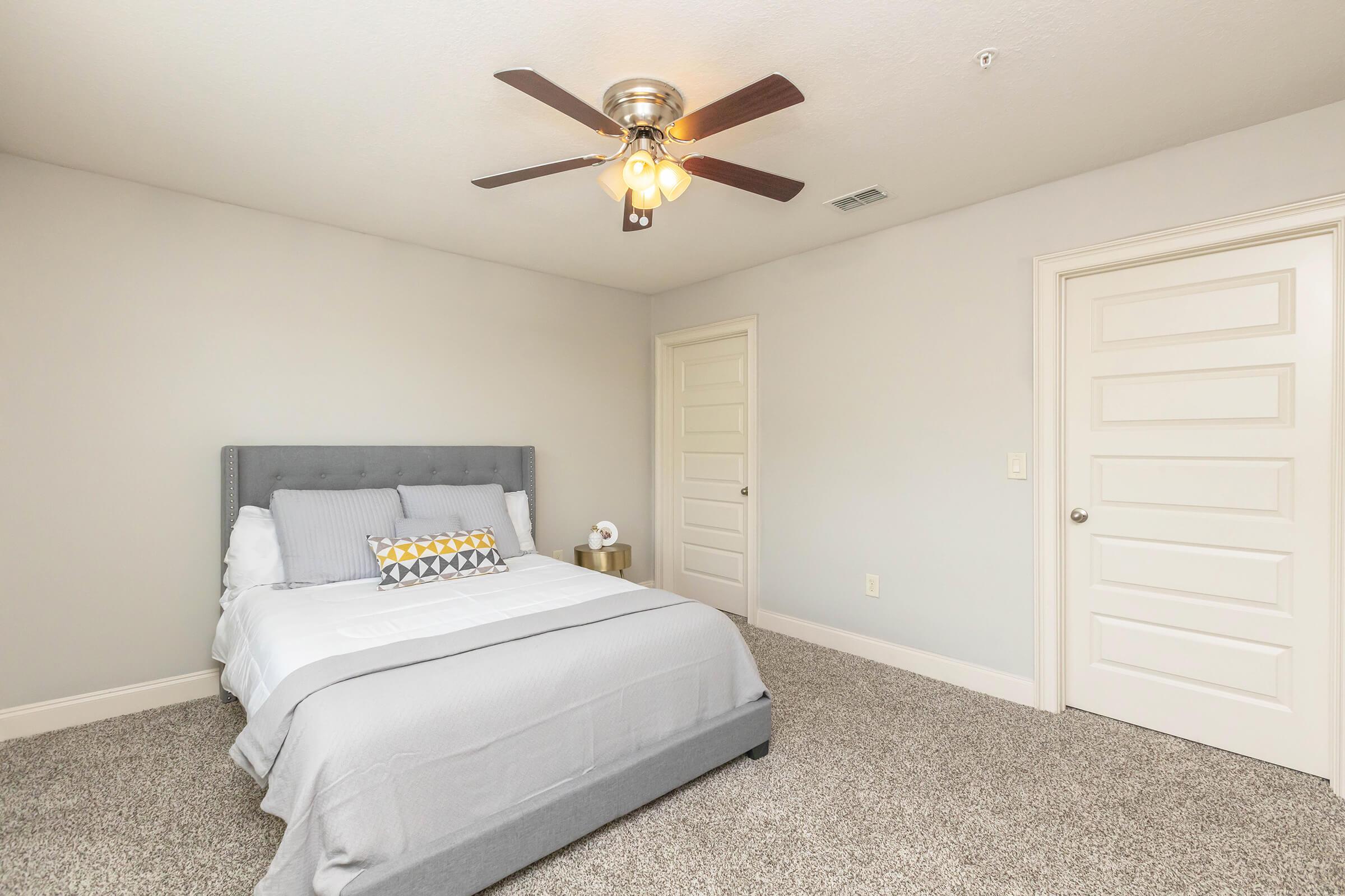 A modern bedroom featuring a gray upholstered bed with white and gray bedding, a decorative yellow accent pillow, and a nightstand. The room has beige carpet, light gray walls, and a ceiling fan with a light fixture. Two white doors are visible, providing access to adjacent rooms.