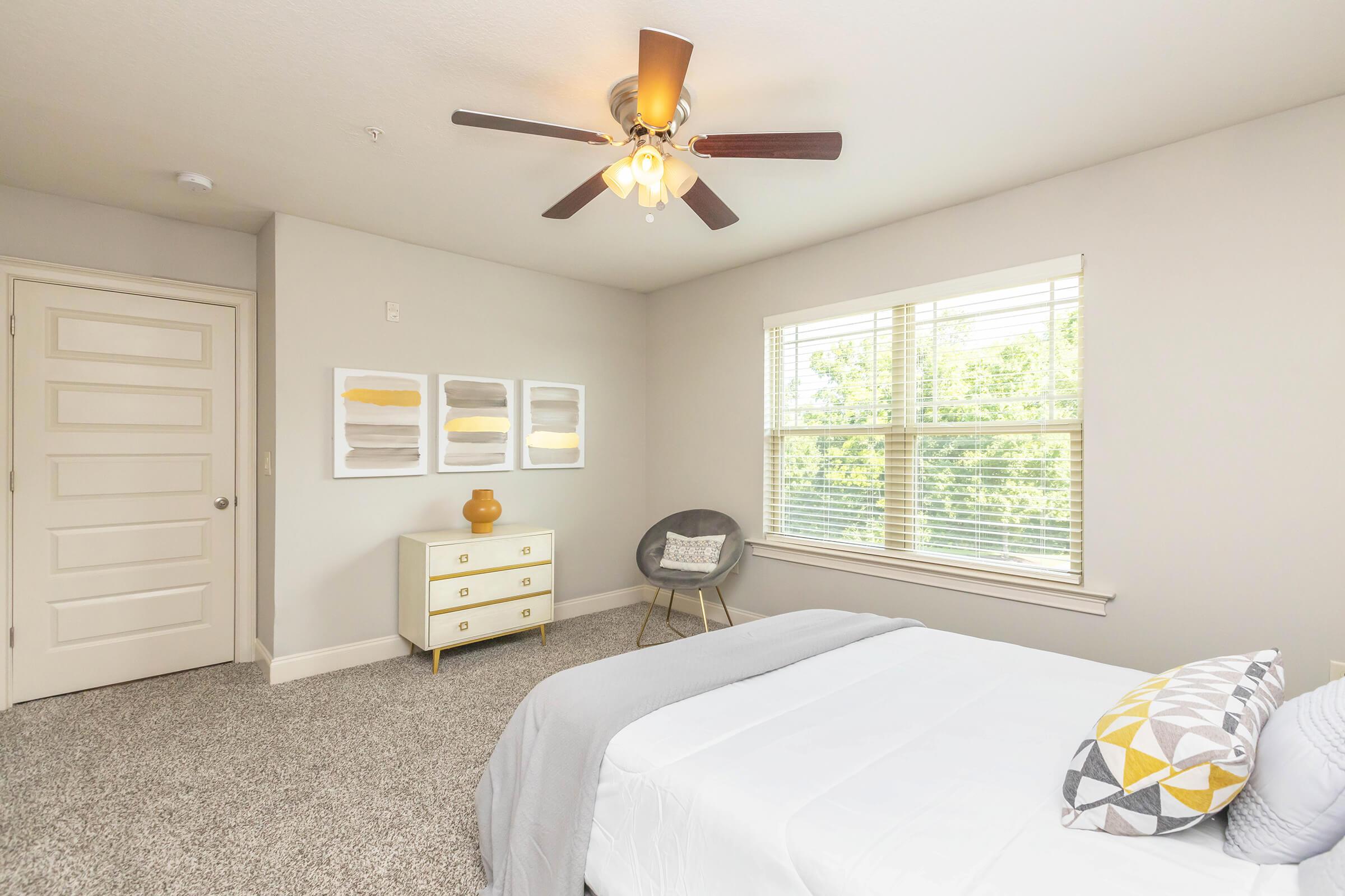 A bright, modern bedroom featuring a white bed with decorative pillows, a light gray blanket, and a stylish chair. The room has a ceiling fan, a large window with natural light, light-colored walls, and art pieces on the wall. A dresser with a decorative vase completes the cozy atmosphere.