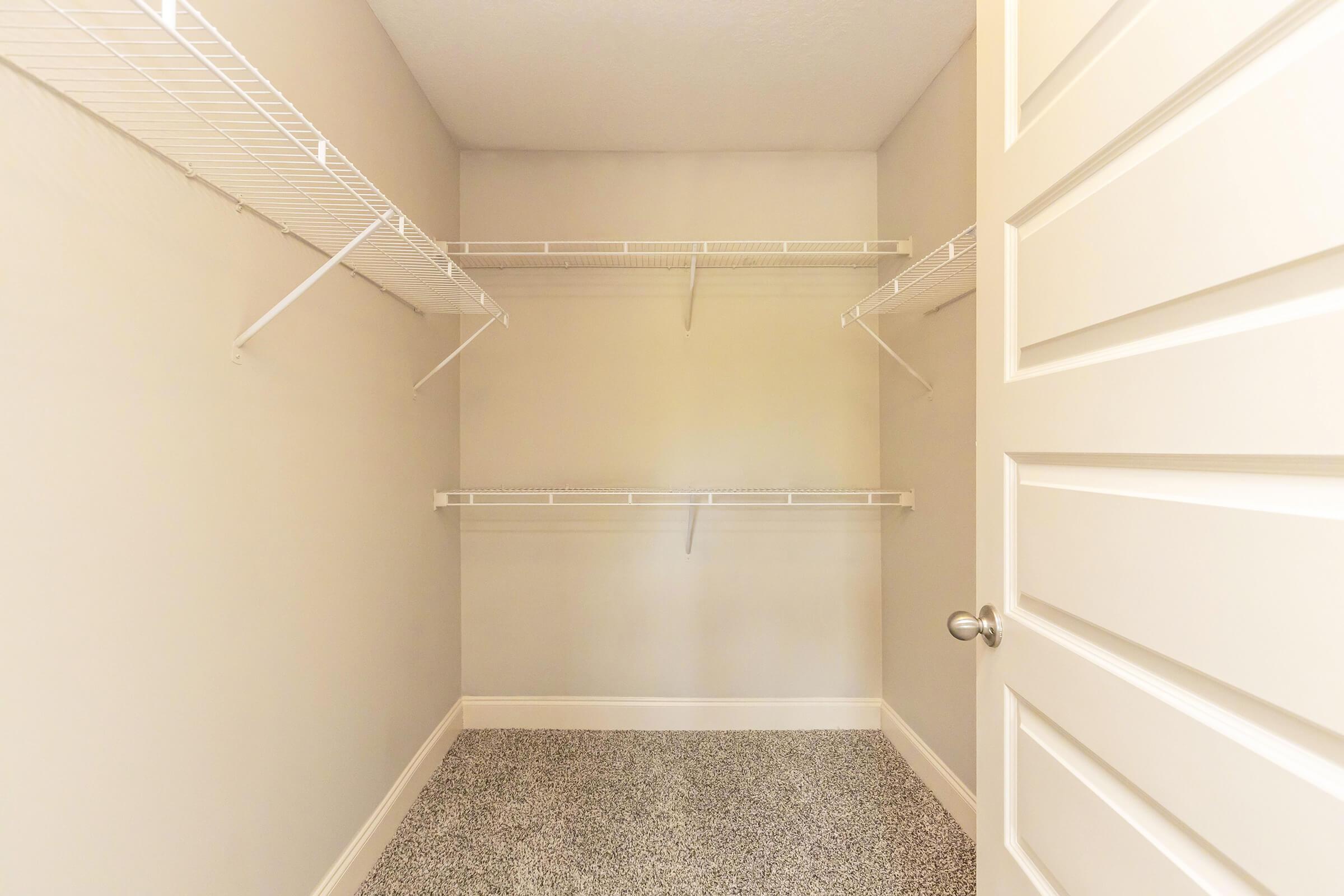 A spacious empty closet with light gray walls and a textured floor. It features two wire shelving units on the side walls and two additional shelves along the back wall. A closed white door with a round metal doorknob is visible on the right side of the image.