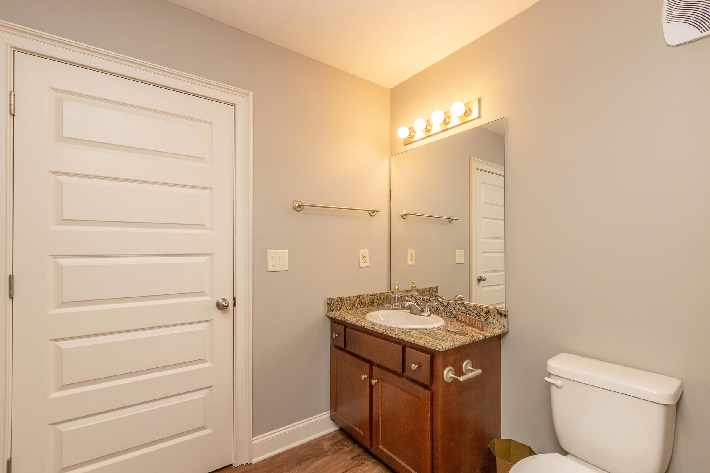 A well-lit bathroom featuring a bathroom vanity with a sink and a mirror above it. There is a wooden cabinet below the sink, a white toilet, and a textured gray wall. A light fixture with multiple bulbs is mounted above the mirror, providing bright illumination. A closed door is visible in the background.