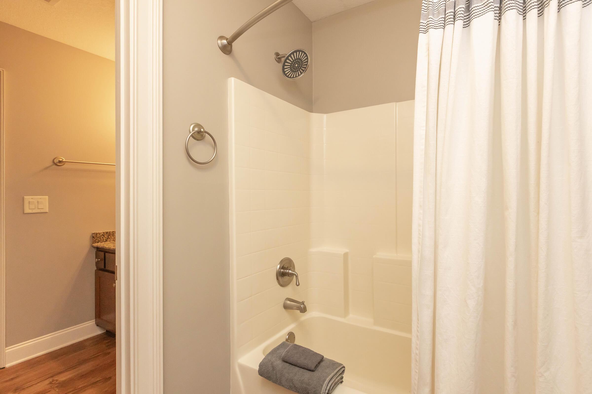 A clean, well-lit bathroom featuring a white bathtub with a shower curtain. The shower area has a modern showerhead and a towel neatly placed on the edge of the tub. The walls are painted in a soft color, and there is a glimpse of a sink area in the background. Wood flooring completes the inviting look.