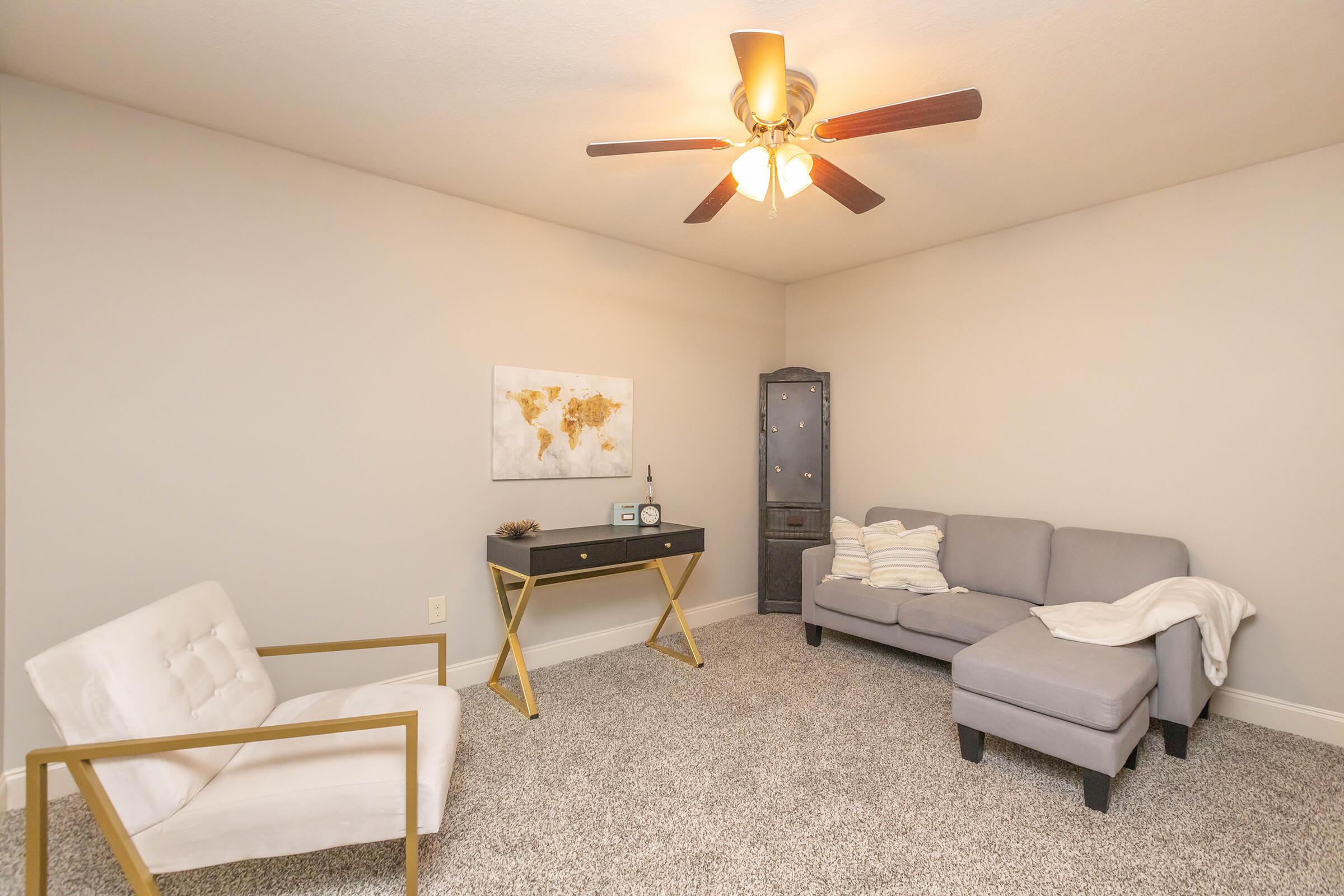 A cozy, minimalistic living room featuring a gray sectional sofa, a white accent chair, a black desk with decorative items, a world map on the wall, and soft carpet flooring. Ceiling fan with light fixture enhances the inviting atmosphere.