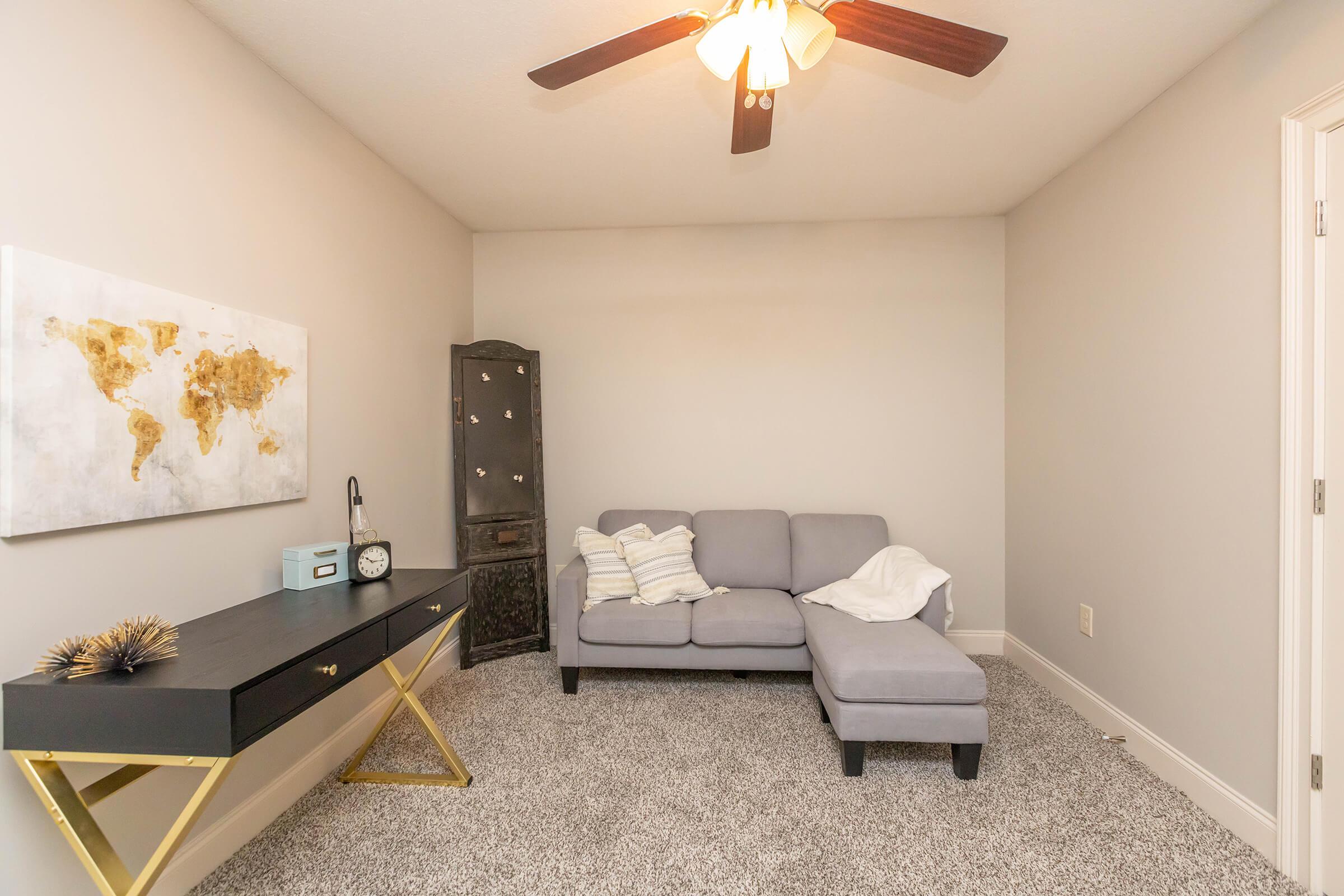 A cozy room featuring a light gray sectional sofa with a throw blanket, a black desk with a clock, and a decorative globe map on the wall. The space has neutral-colored walls and soft carpet, with a vintage-looking cabinet in one corner and a ceiling fan providing light and ventilation.