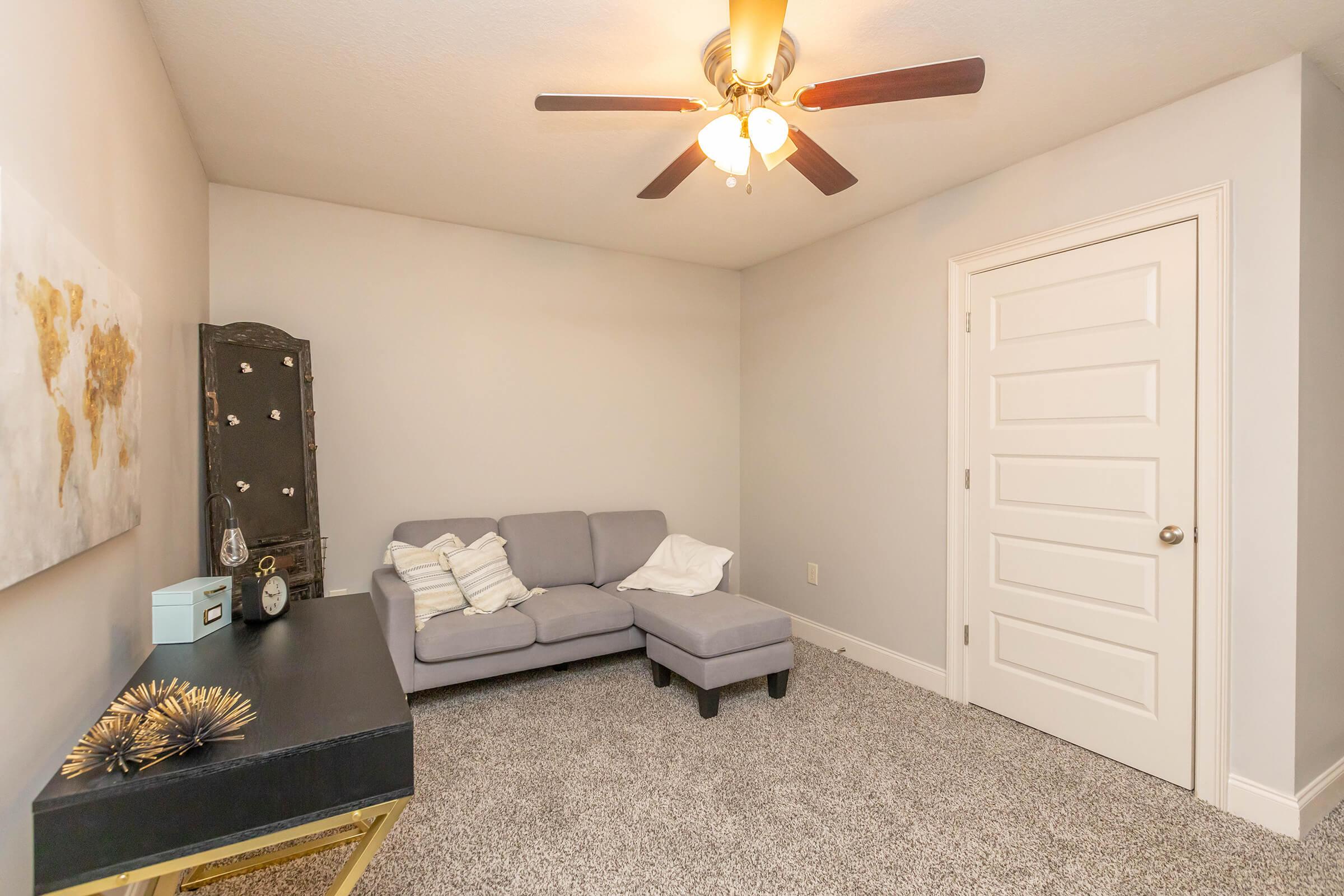 A cozy, minimalist room featuring a gray sectional sofa with a white throw blanket, a small black desk with decorative items, a wall clock, and a world map artwork. The space has a ceiling fan and a closed white door, with light-colored walls and plush carpet flooring, creating a warm and inviting atmosphere.