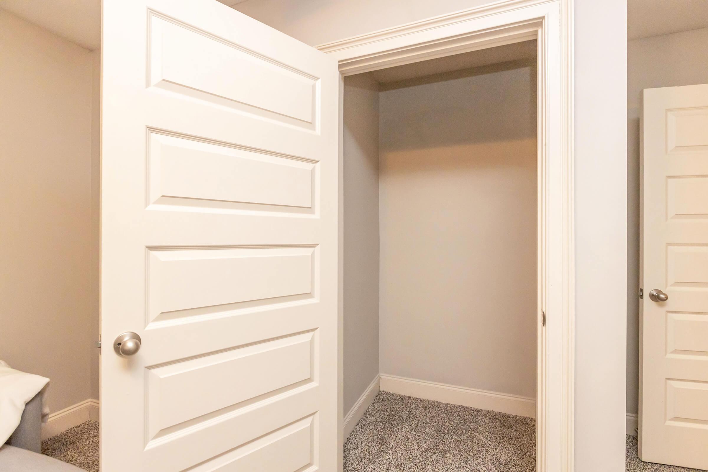 A view of a small, empty closet with a white, paneled door partially open. The walls are painted a light color, and the floor is covered with gray carpet. Another closed door is visible in the background, leading to a different room.