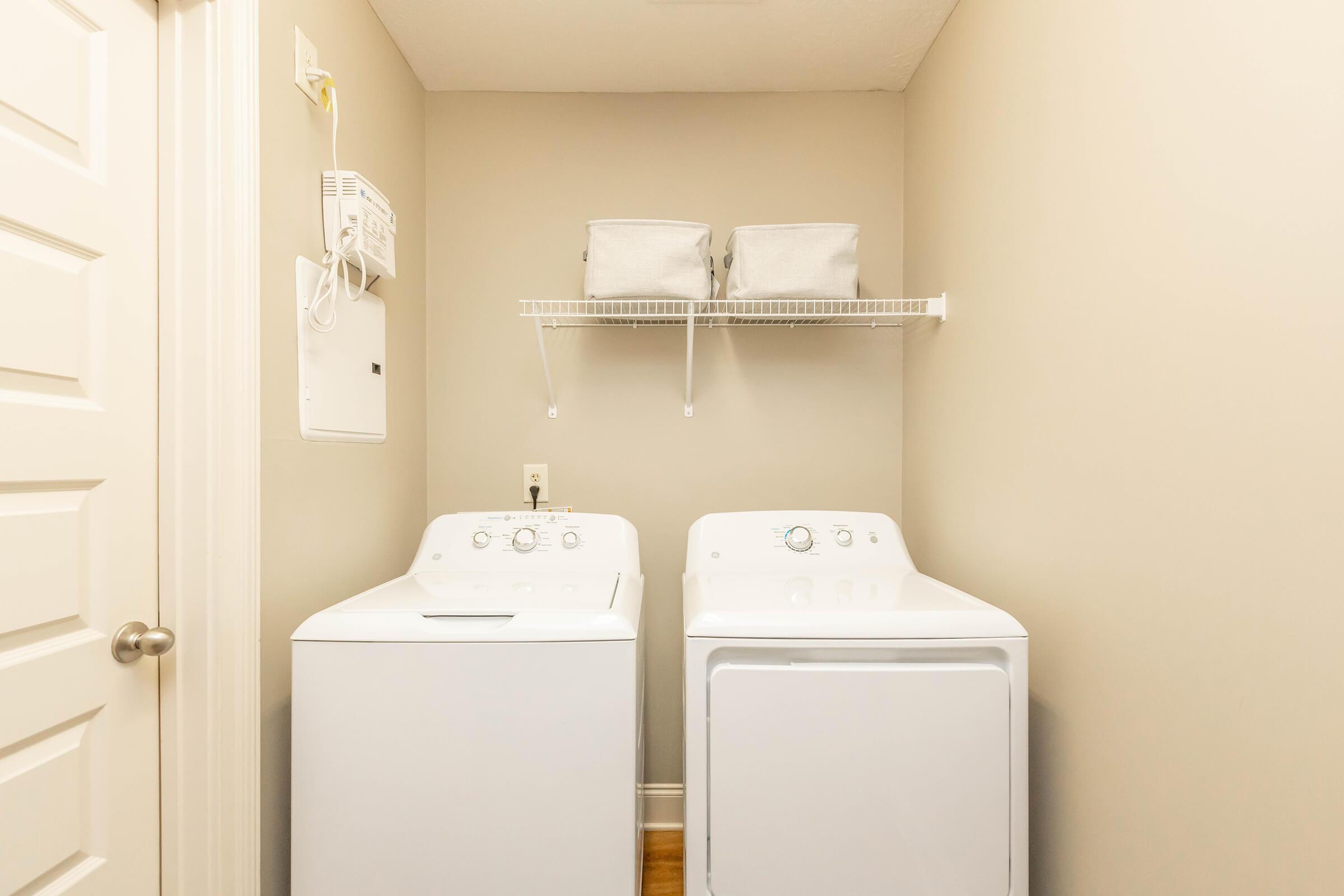 A laundry room featuring a white washing machine and a white dryer side by side. Above them, there is a shelf with two storage bins. The walls are painted a light color, and there's a closed door on the left. The space is clean and organized, showcasing a practical laundry setup.