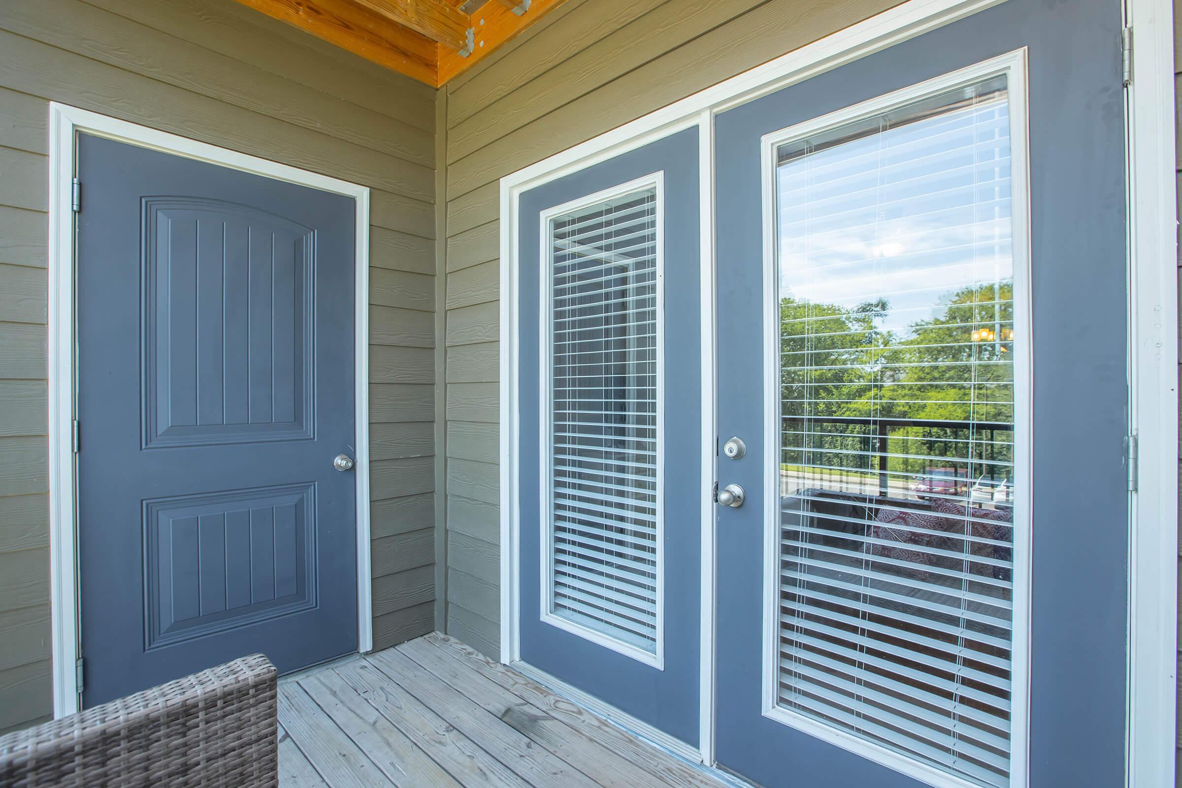 Two gray doors on a wooden deck, one solid and one with large glass panels featuring blinds. The doors are framed by a light-colored wall, and outside, greenery can be seen through the glass. There's a wicker chair in the foreground, adding to the outdoor living space feel.