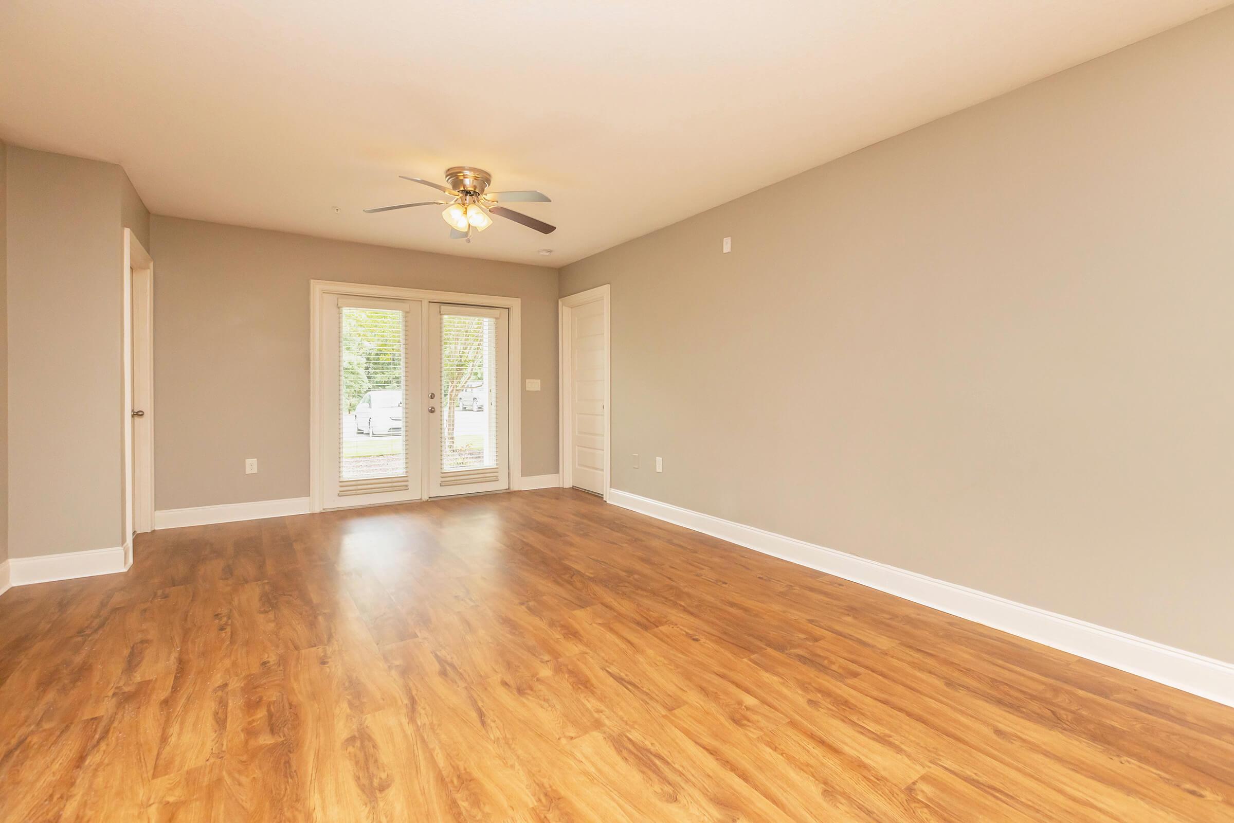 A spacious, empty room with light brown wooden flooring and light gray walls. There is a ceiling fan with three light bulbs and a set of double doors leading to an outdoor area. The room has a modern, minimalistic design and a single white door on the left side.