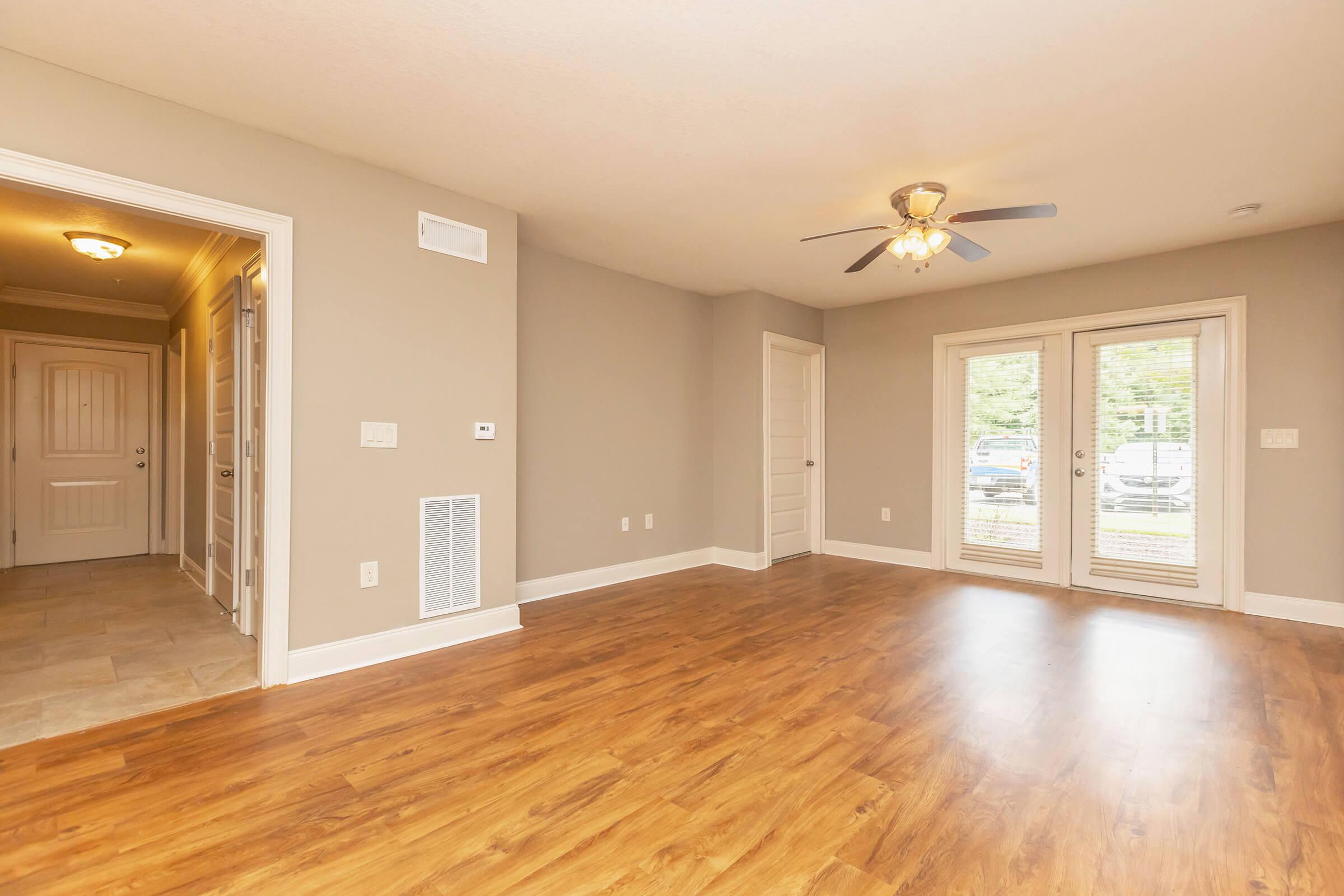 A spacious living room with light wood flooring, a ceiling fan, and large sliding doors leading to an outdoor space. The room features neutral-colored walls and is connected to a hallway with white doors. Natural light brightens the area, creating a welcoming atmosphere.