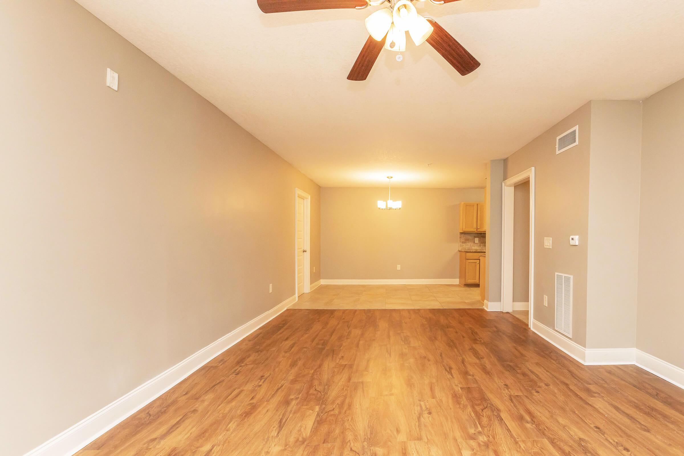 A spacious room with light brown wooden flooring and a ceiling fan. The walls are painted in a soft gray color. On the left, there's a doorway leading to another room, and in the back, an open kitchen area with wooden cabinets is visible. Natural light illuminates the space.