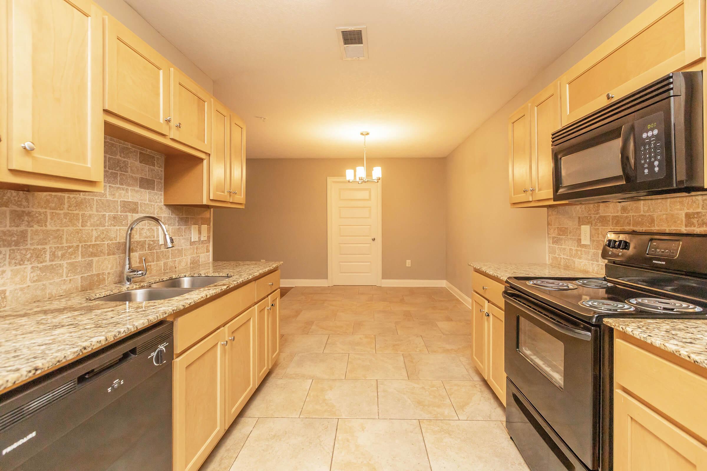 A modern kitchen featuring light wooden cabinets, granite countertops, a double sink, and stainless steel appliances. The floor is tiled, and there is a dining area with a chandelier visible in the background. The walls are painted a light neutral color, enhancing the spacious feel of the room.