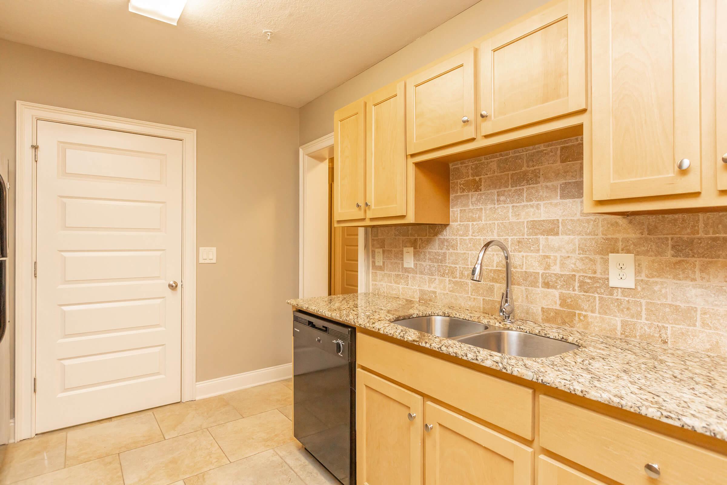 A modern kitchen featuring light wooden cabinetry, a double sink, a granite countertop, and a stainless steel dishwasher. The backsplash consists of beige tiles, and there's a white door leading to another room in the background. Natural light illuminates the space, creating a warm atmosphere.