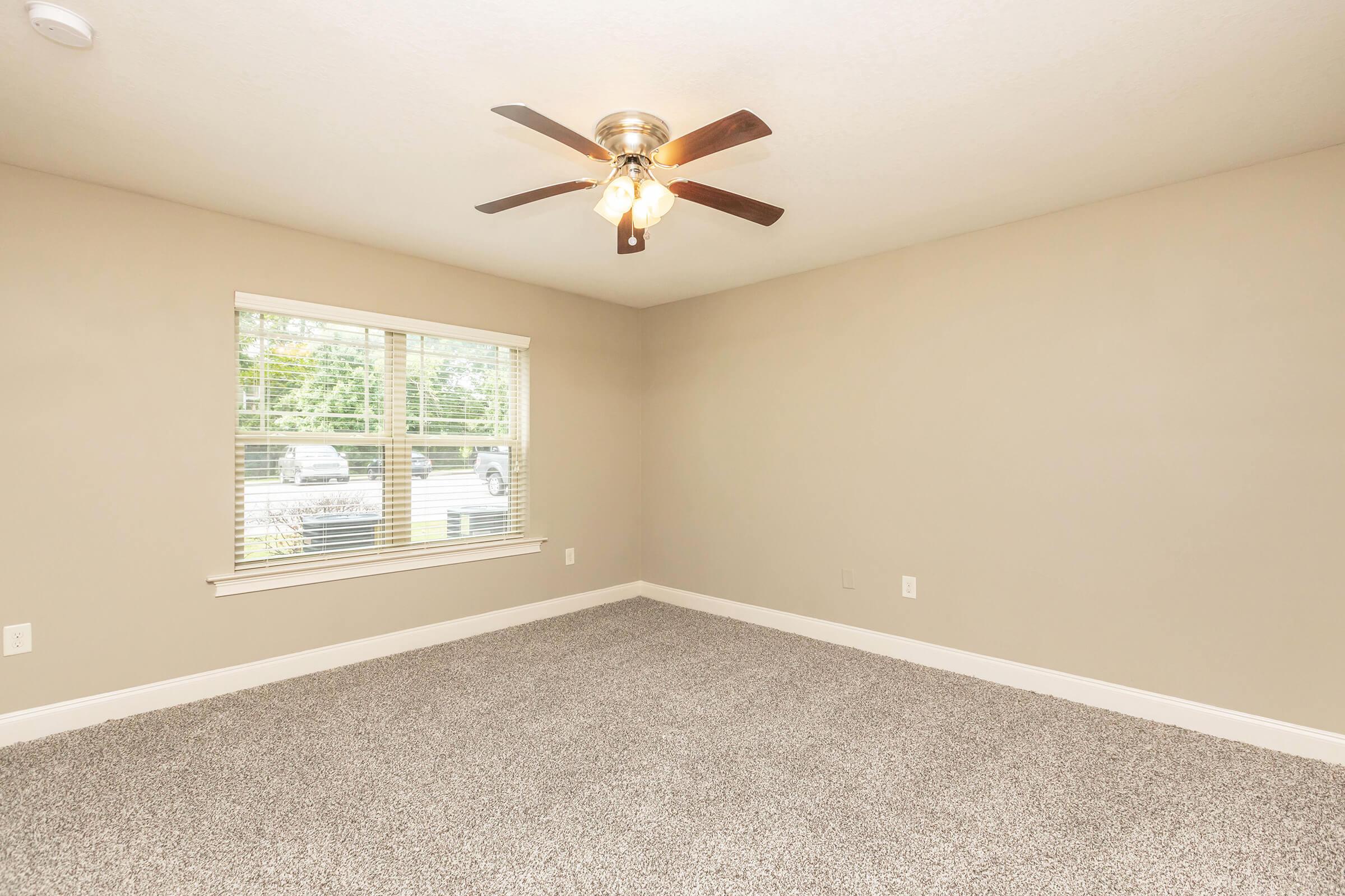 A well-lit, empty room with beige walls and a ceiling fan. The floor is covered in light gray carpet. A large window with blinds allows natural light to enter, revealing a view of the outdoors. The space is clean and ready for furnishings, providing a blank canvas for personal decoration.