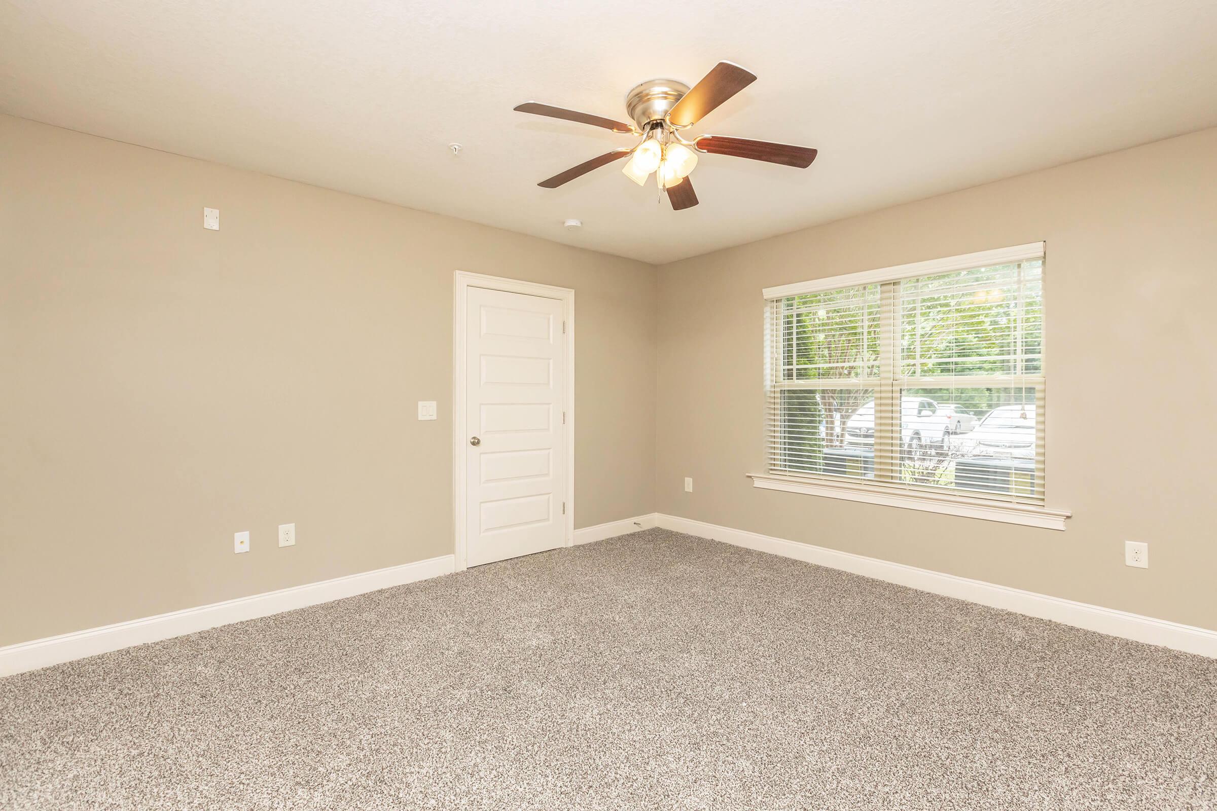 A spacious, empty room featuring light gray carpet, a ceiling fan with three lights, and a window with blinds overlooking an outdoor area. The walls are painted a soft beige, and there's a white panel door leading to another space. Natural light fills the room, creating a bright atmosphere.