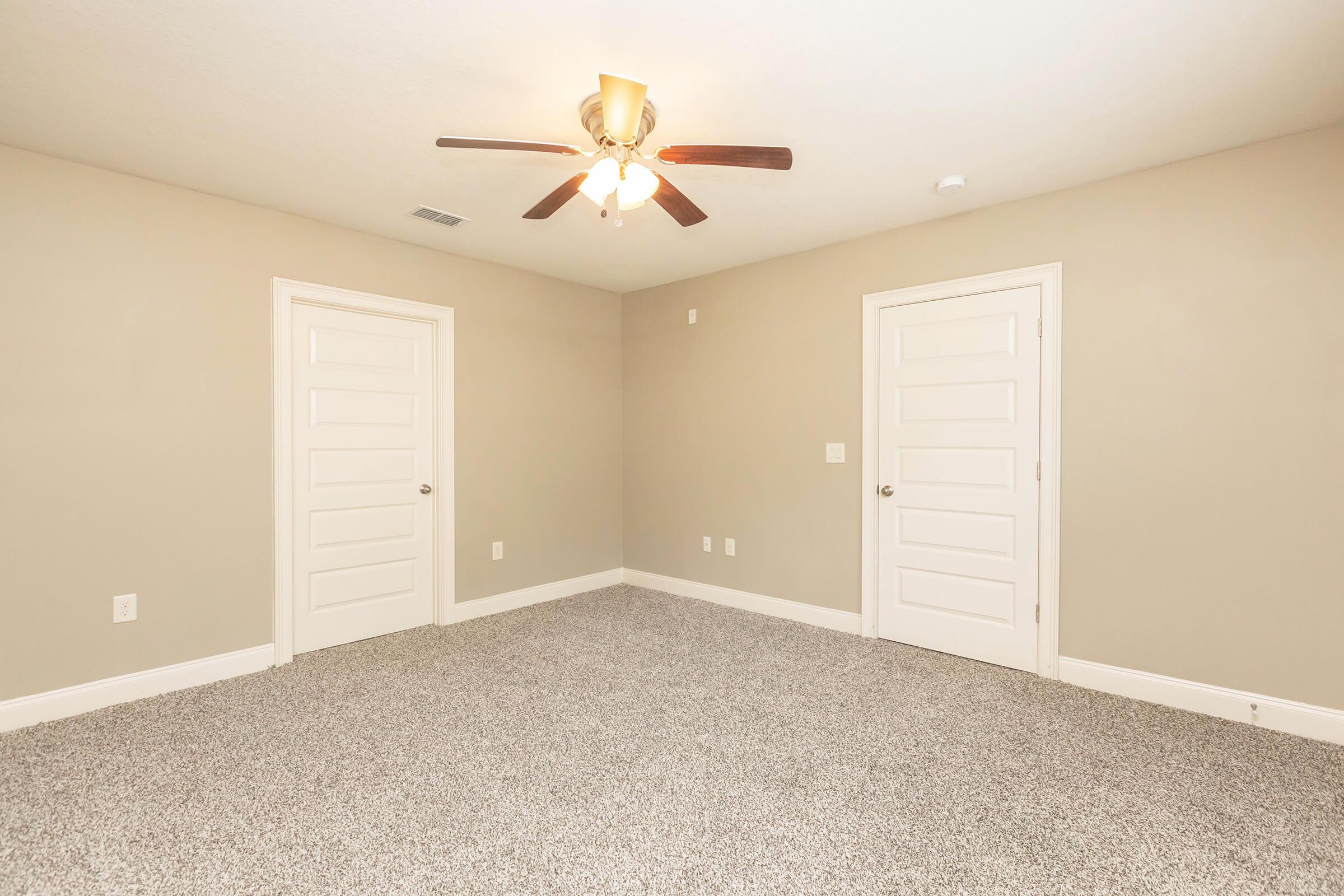 A spacious, empty room featuring light beige walls and a ceiling fan with wooden blades. The floor is covered in plush carpet, and there are two white paneled doors on opposite walls. The overall ambiance is bright and inviting, suitable for various purposes such as a bedroom or office.