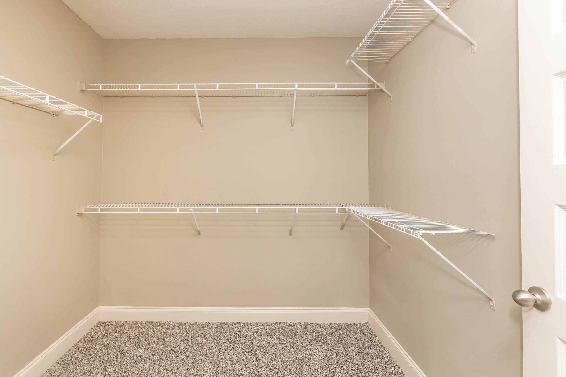 An empty closet with beige walls and gray speckled flooring. Wire shelving is installed along the walls, with several shelves positioned at various heights, providing storage space. The door is partially closed, showing a handle and part of the door frame. The overall appearance is clean and minimalistic.