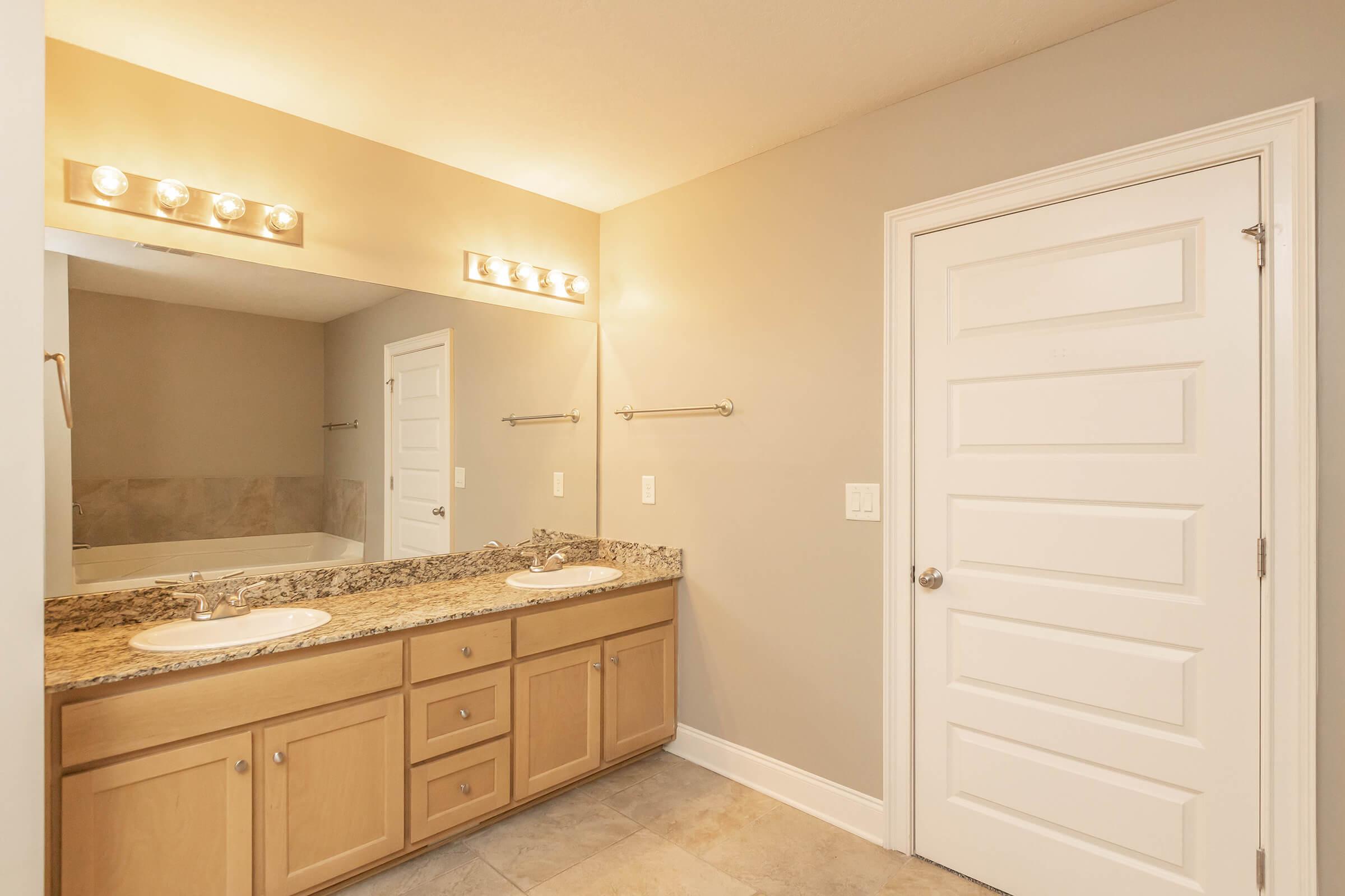 A well-lit bathroom featuring a double sink vanity with granite countertops, a large mirror above, and a spacious layout. The walls are painted a neutral color, and there is a white door leading to another room. The flooring is tile, giving the space a clean and modern appearance.