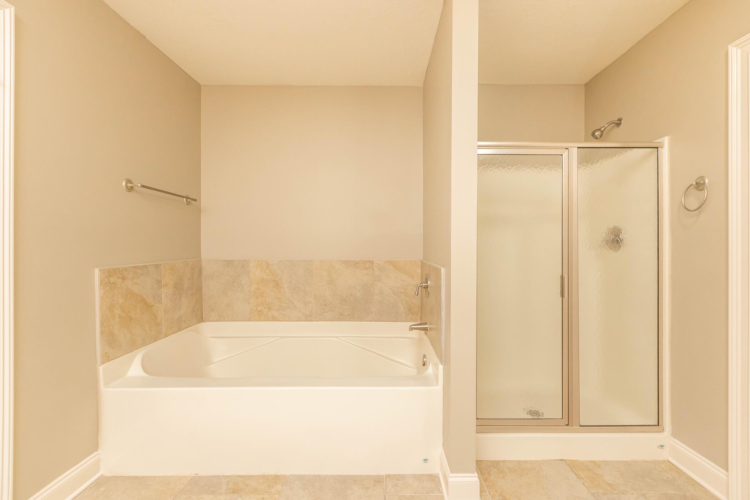 A modern bathroom featuring a large white bathtub, a glass-enclosed shower, and light beige walls. The floor is tiled with a subtle pattern, and there are stainless steel fixtures. The overall aesthetic is clean and spacious, creating a serene atmosphere.