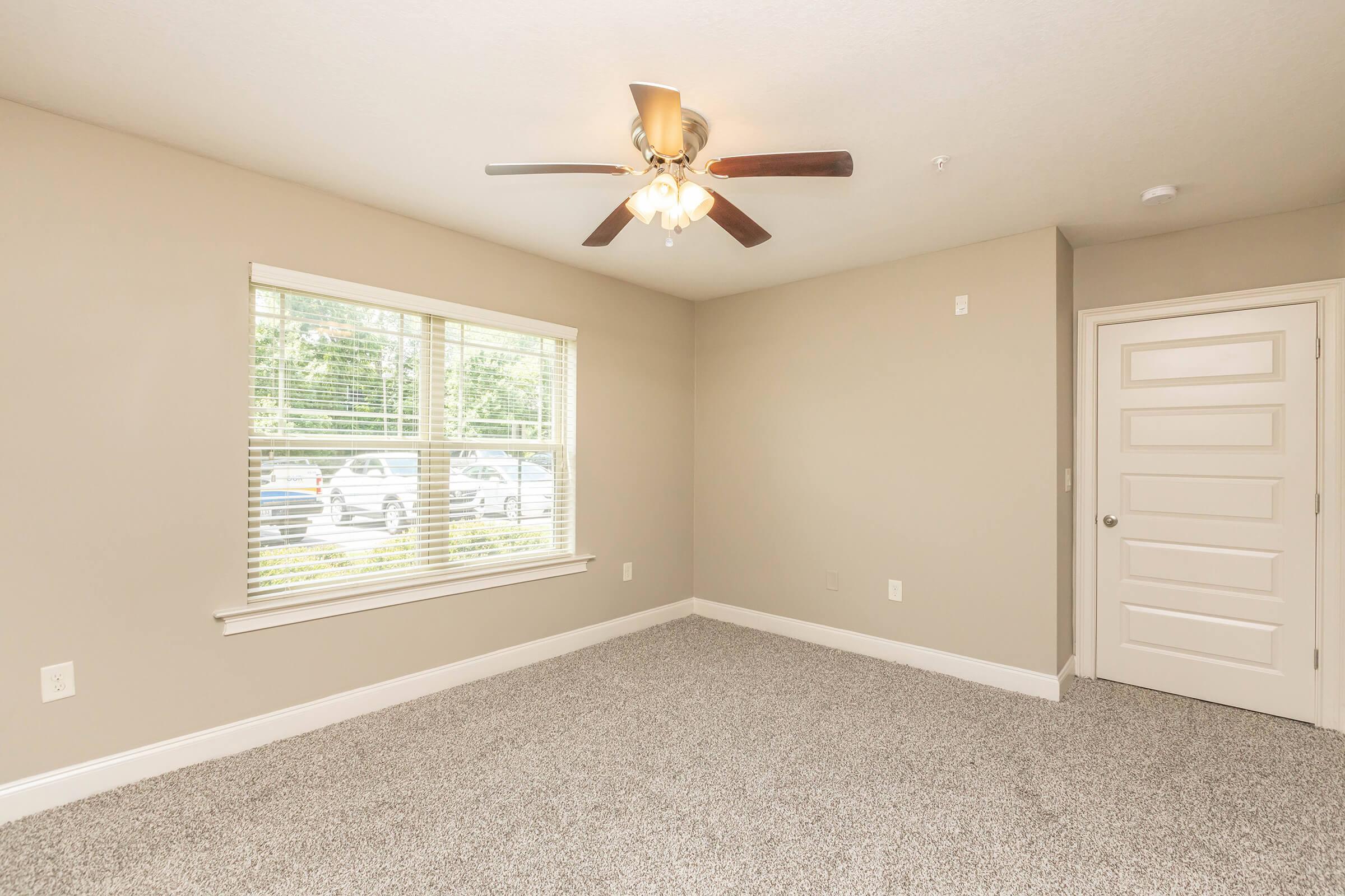 A well-lit, empty room with beige walls and soft gray carpeting. There's a ceiling fan with light fixtures and a large window allowing natural light to flood in. The door is white with a simple design, and the overall space appears clean and modern, suitable for various用途.