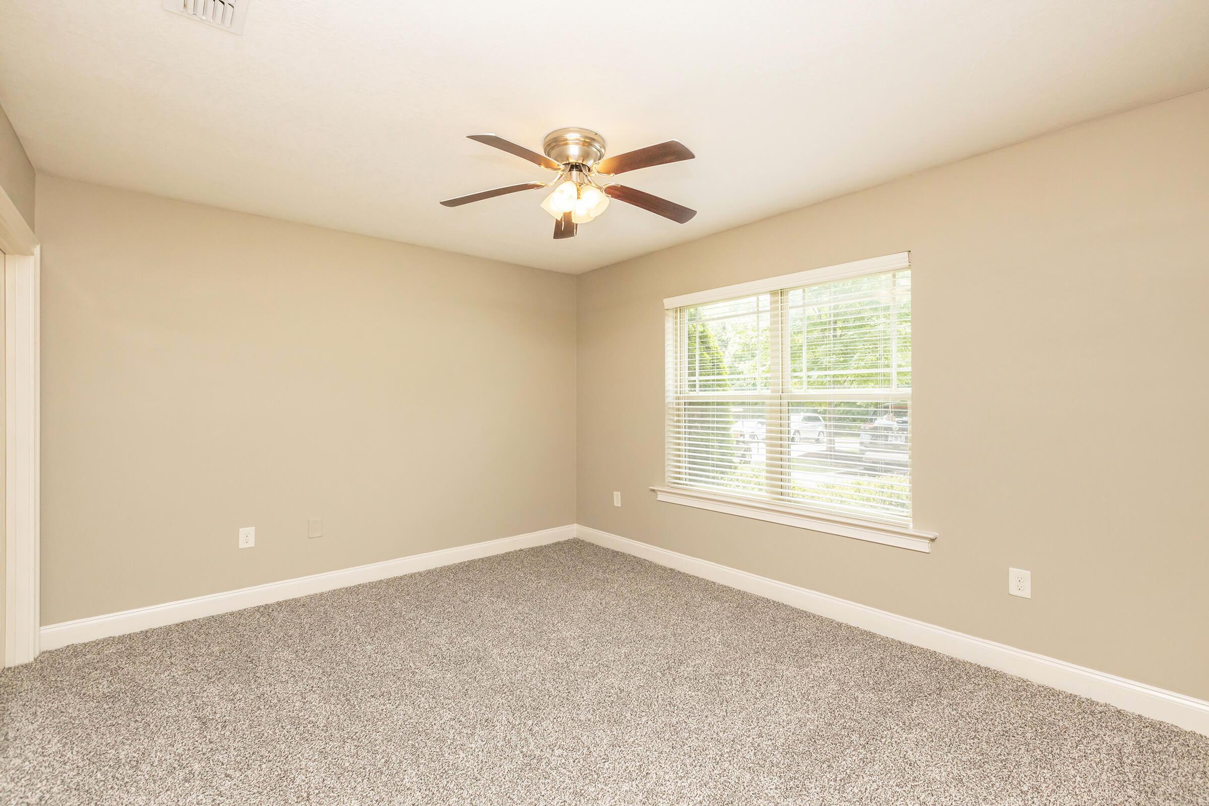 A spacious room with light beige walls and a ceiling fan. The floor is covered in gray carpet. There is a large window with white blinds that lets in natural light, offering a view of the outside. The room has no furniture, creating a blank canvas for decoration.