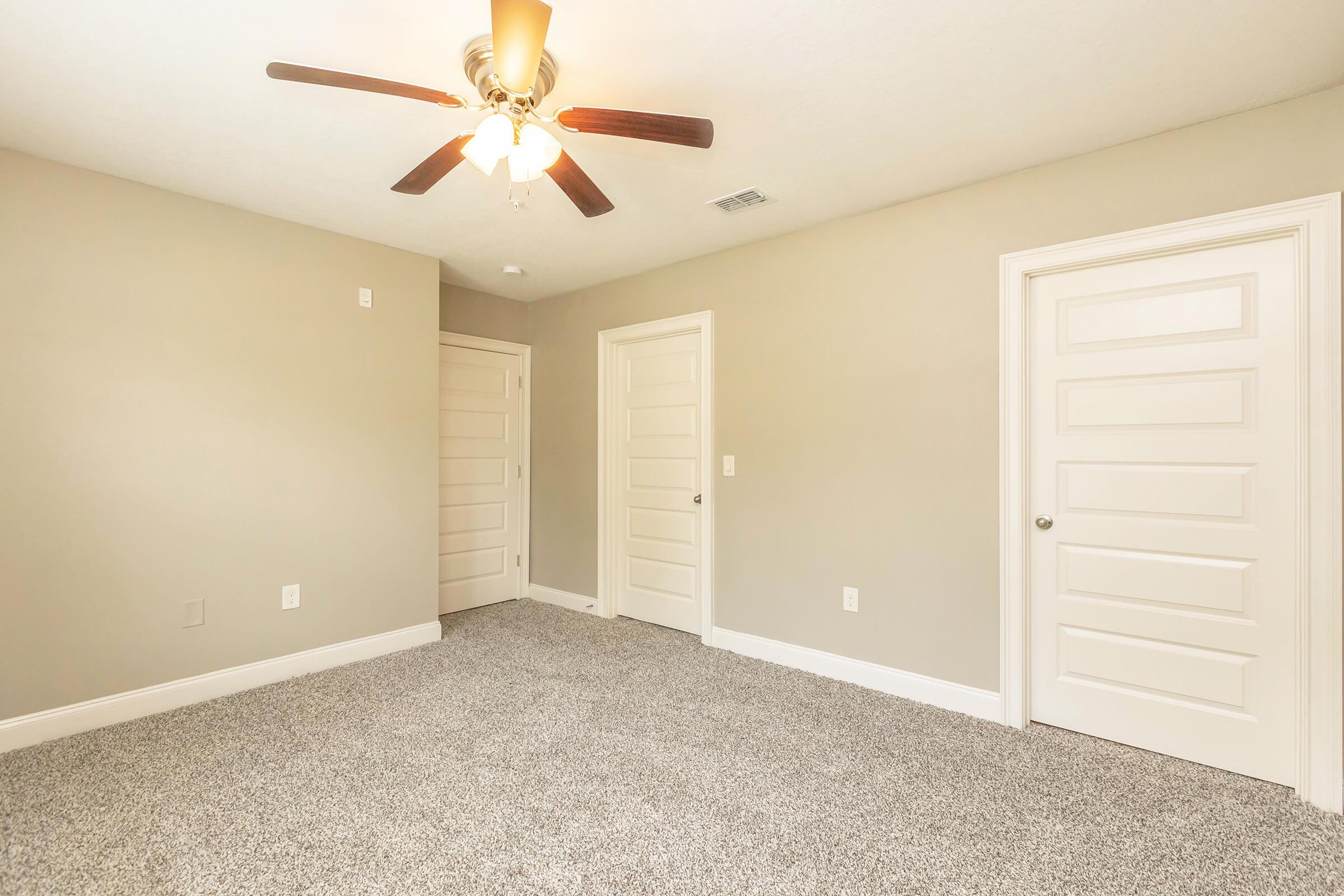 A spacious, empty bedroom featuring light gray walls, a ceiling fan with wooden blades, and plush carpet flooring. Two white doors are visible, indicating closet space and entry to another room. The room is well-lit, creating a calm and inviting atmosphere.