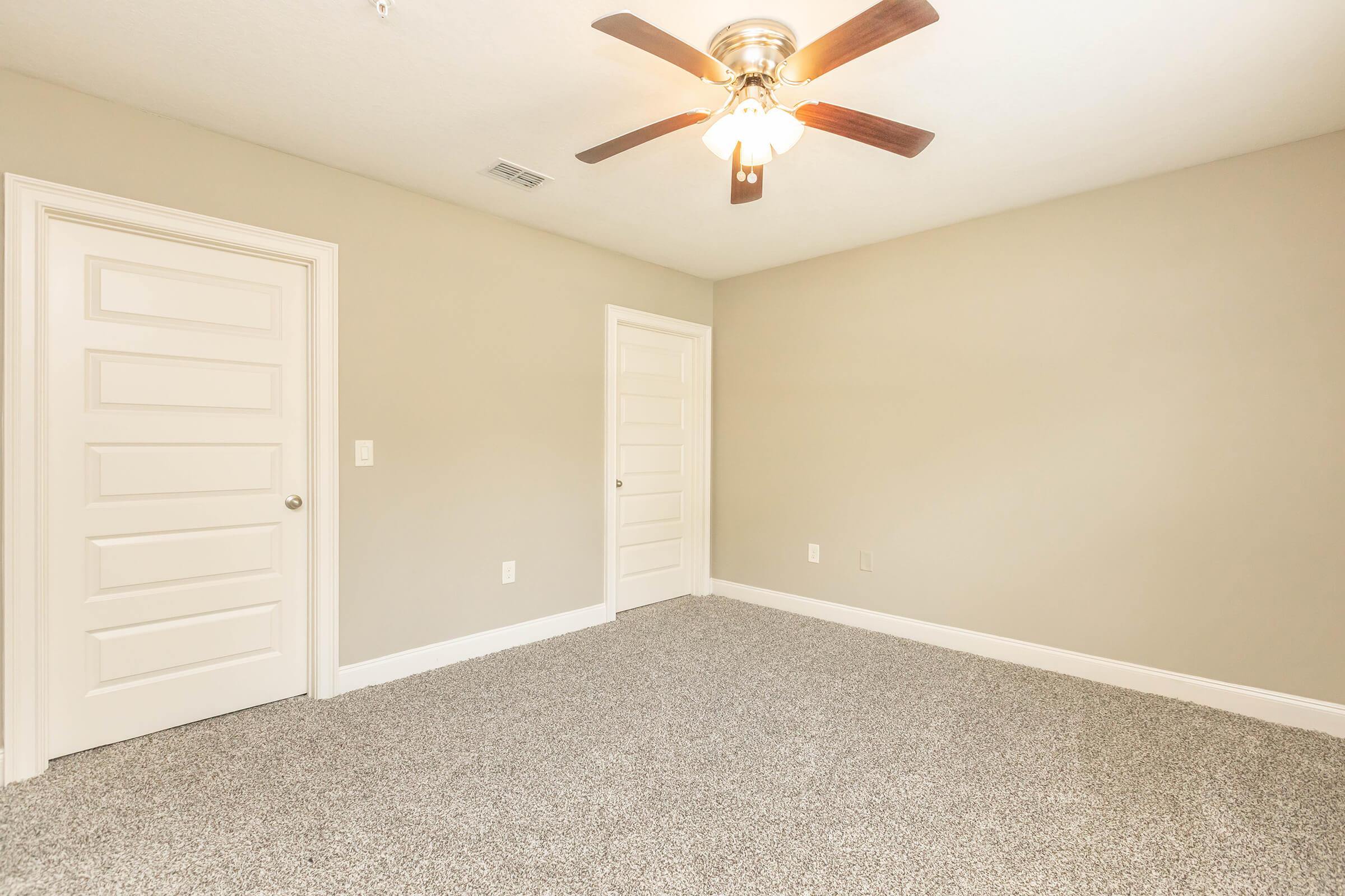 A well-lit, empty bedroom featuring soft gray carpeting, light beige walls, and a ceiling fan. Two closed doors are visible, one leading to a closet. The space appears clean and ready for furnishing, with ample natural light highlighting the room's neutral color scheme.