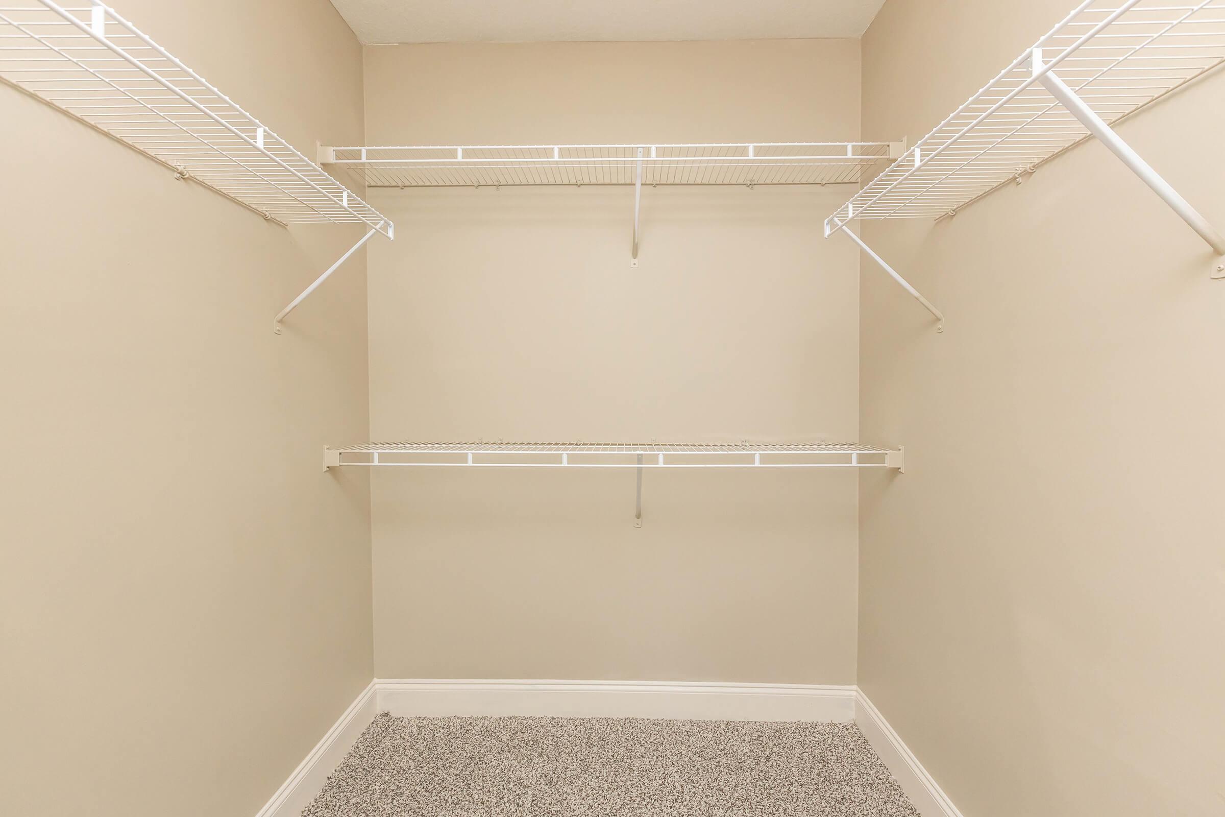 A vacant closet with beige walls and a carpeted floor, featuring two wire shelves along the side walls and one shelf above, all in white. The space is empty and well-lit, creating a clean and organized appearance.