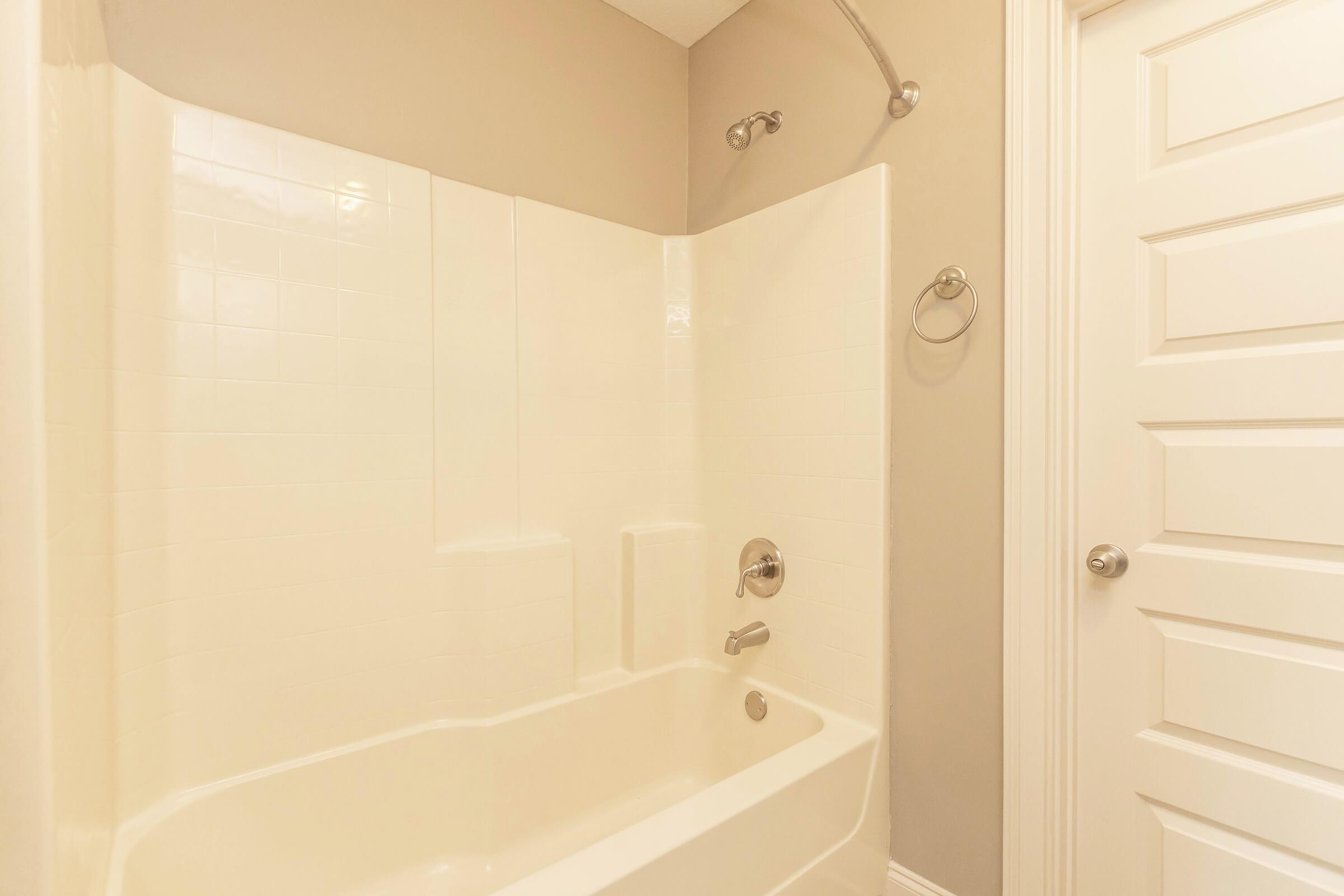 A clean, modern bathroom featuring a white tub and shower combination. The walls and tub have a smooth, glossy finish. A chrome showerhead and faucet are visible, along with a towel ring and a closed door with sleek hardware. The overall decor is simple and neutral, creating a calm atmosphere.