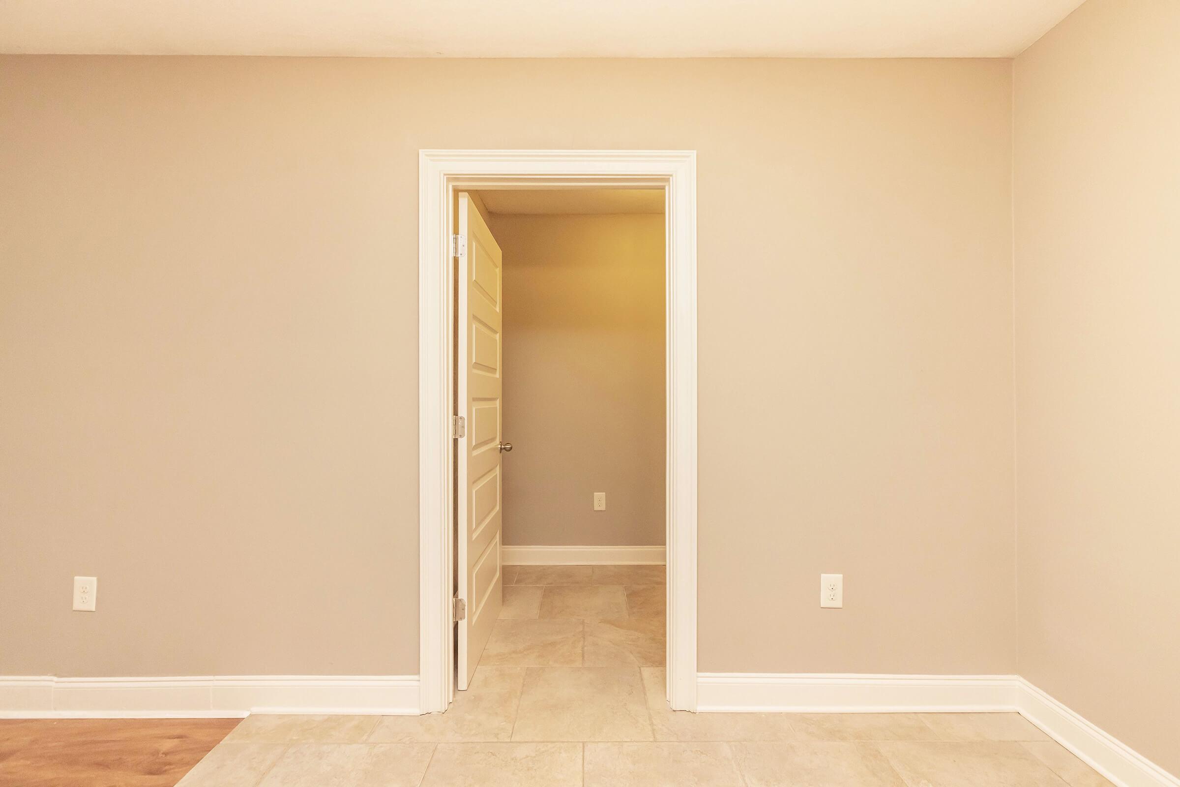 A simple, empty room with light gray walls and a tiled floor. A white door is slightly ajar, leading to another space. The lighting is soft and subdued, creating a neutral and inviting atmosphere. No furniture or decorations are present, emphasizing the spacious and minimalistic design.