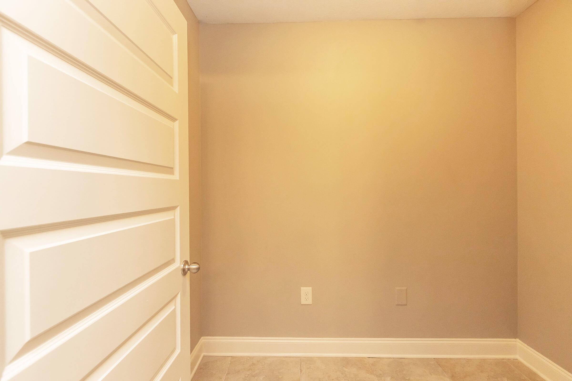 A clean, minimalistic interior of a small room with beige walls and tile flooring, featuring a closed white door on the left and a blank wall on the right. The lighting is soft and neutral, creating a calm atmosphere.