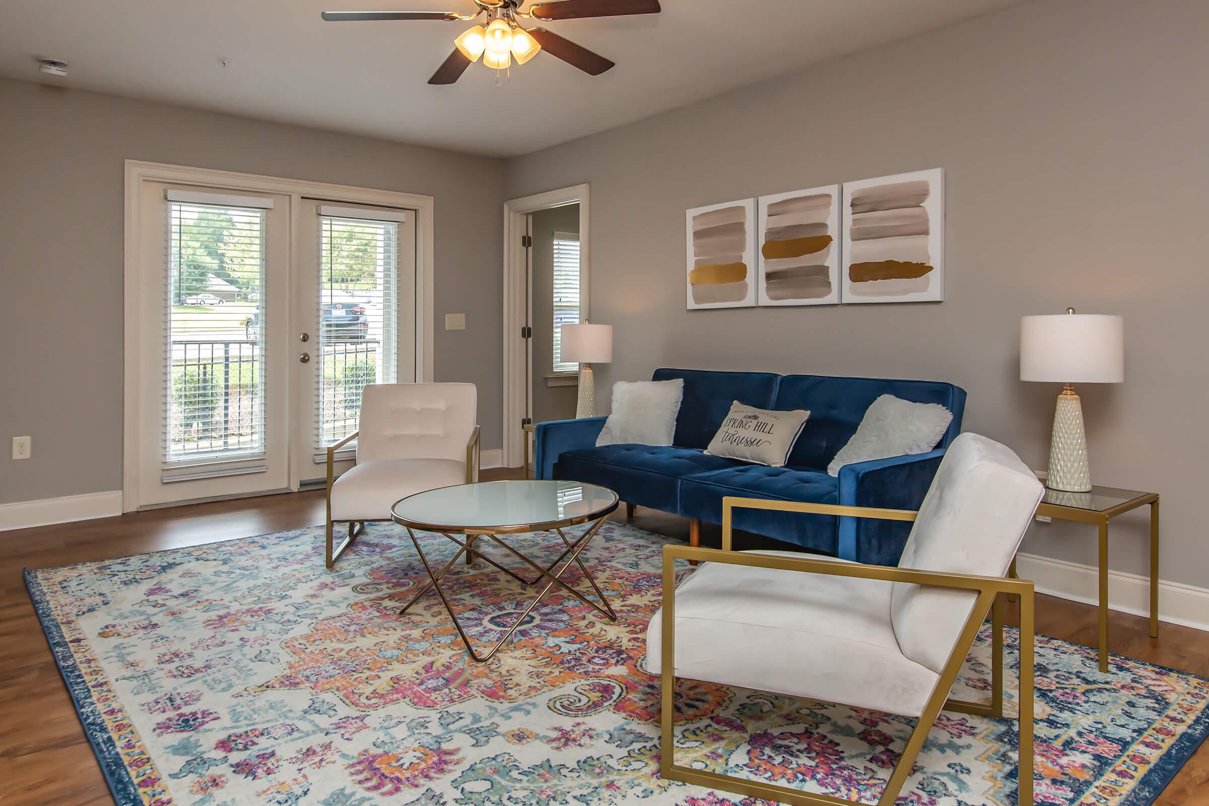 A cozy living room featuring a blue velvet sofa, two white accent chairs, a glass coffee table, and decorative pillows. The space is brightened by natural light coming through sliding glass doors and is complemented by a colorful area rug and modern wall art.