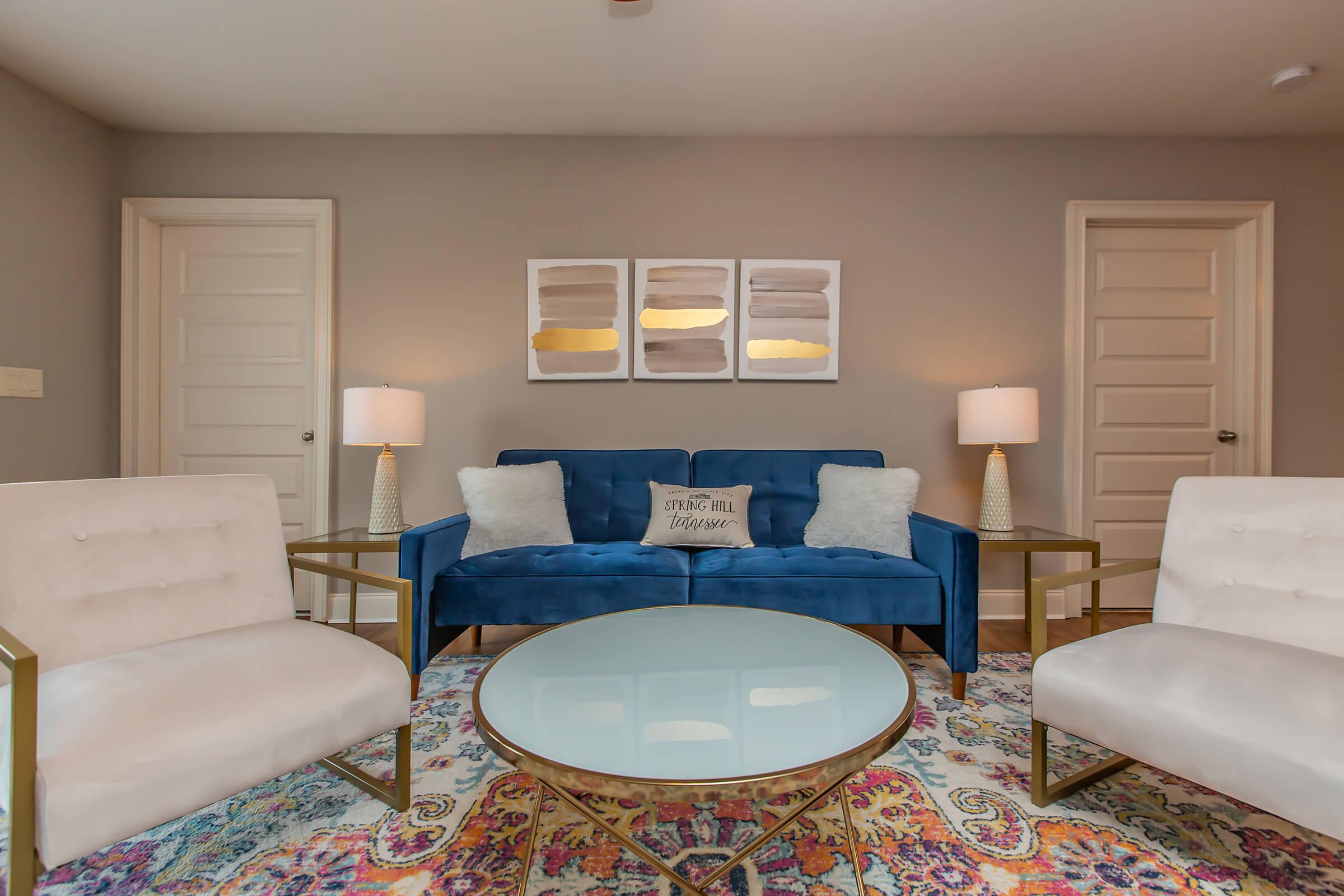 A cozy living room featuring a blue velvet sofa with two decorative pillows, flanked by two modern white chairs. A round glass coffee table sits in the center on a colorful patterned rug. Two lamps provide soft lighting, and three abstract art pieces are mounted on the wall above the sofa. Two closed doors are visible in the background.