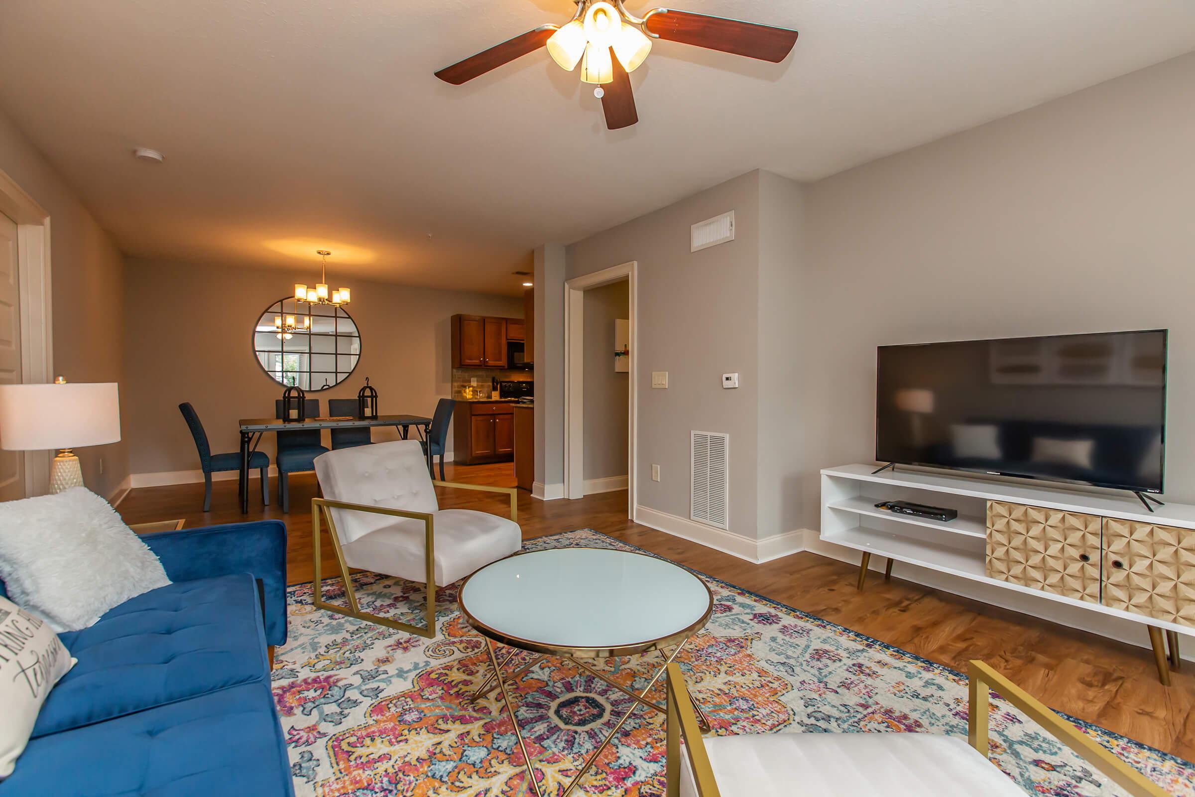 A cozy living room featuring a blue sofa, gold-accented chairs, and a round coffee table on a colorful area rug. A flat-screen TV is on a modern white entertainment unit. In the background, a dining area with a round table and stylish light fixture is visible, creating a warm and inviting atmosphere.