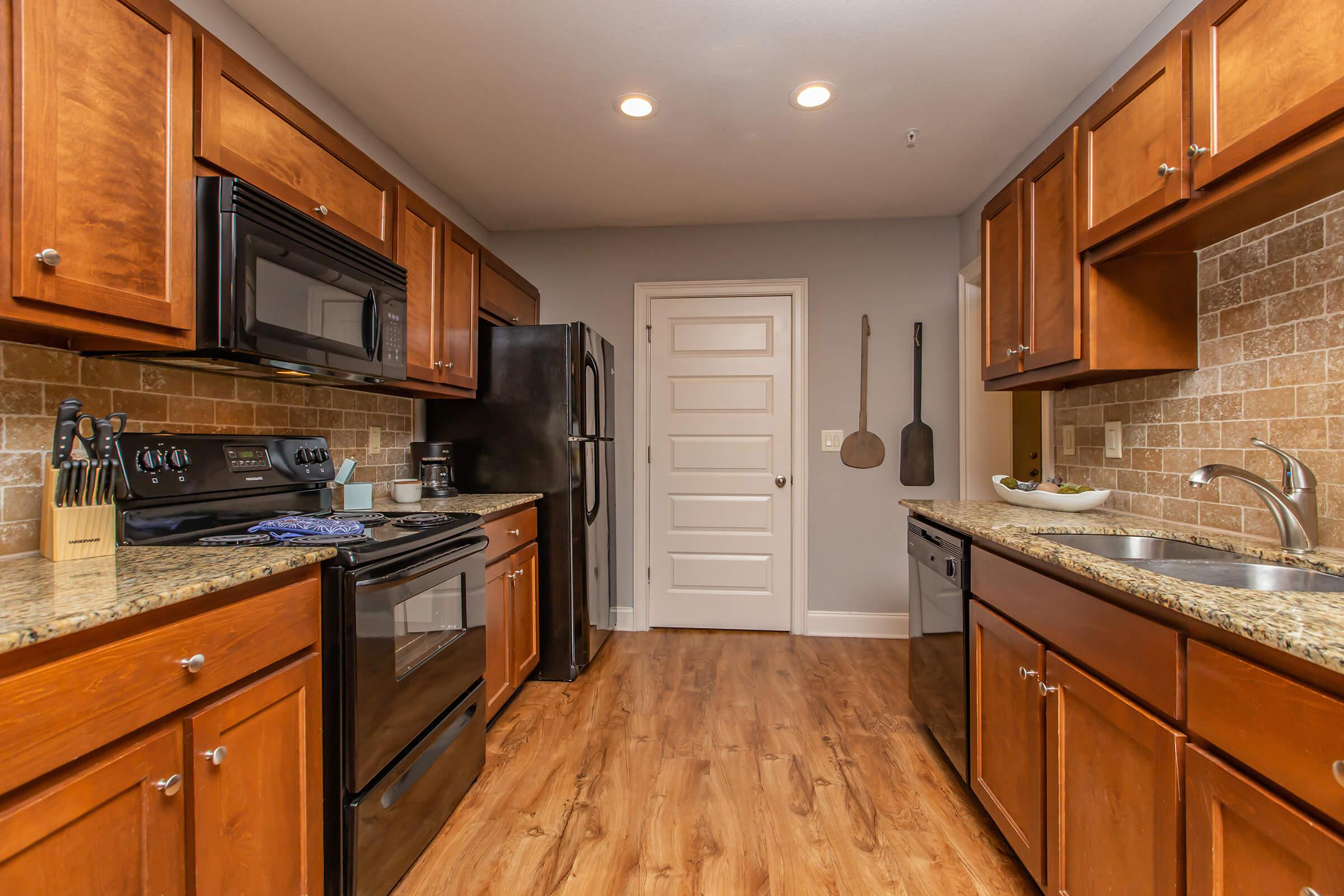 A modern kitchen featuring dark wood cabinets, granite countertops, and stainless steel appliances. It includes a black stove, microwave, and refrigerator, with a dual sink. The walls are a neutral color, and there is a door leading to another room. Wooden utensils hang on the wall, adding a rustic touch.