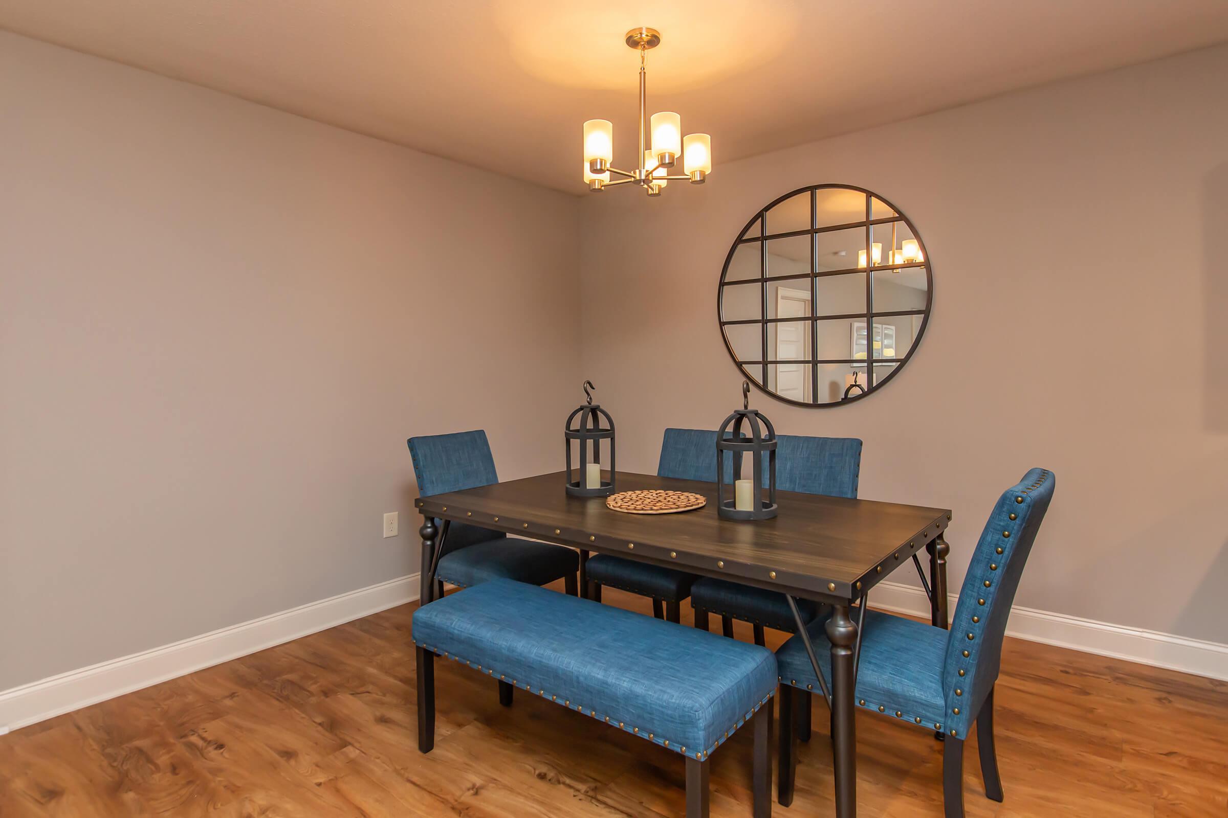 A modern dining room featuring a rectangular wooden table with a dark finish, surrounded by four blue upholstered chairs and a bench. A round mirror hangs on the wall, reflecting the light from a stylish ceiling fixture. The floor is hardwood, adding warmth to the space.