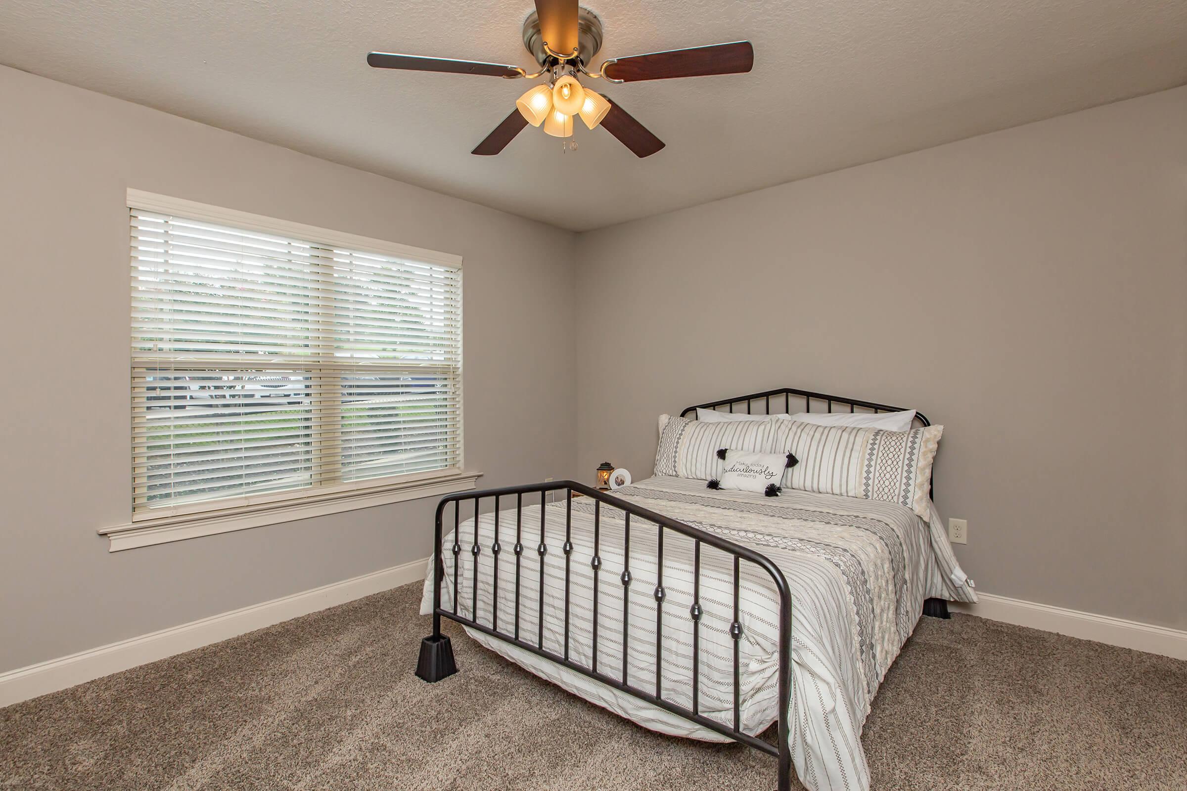 A cozy bedroom featuring a metal bed frame with white and striped bedding. The room has light gray walls and a carpeted floor. A ceiling fan with lights hangs above, and a window with horizontal blinds allows natural light to enter, creating a bright and inviting atmosphere.