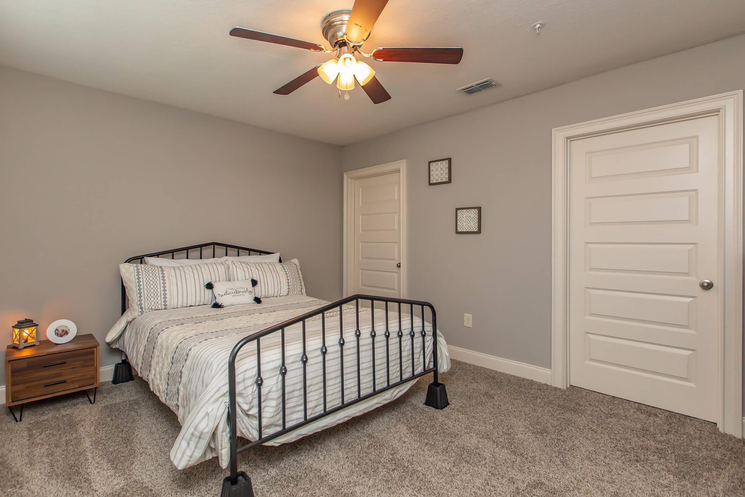 A cozy bedroom featuring a metal bed frame with a striped comforter and decorative pillows. There is a nightstand with a lamp next to the bed, light gray walls, plush carpet, and a ceiling fan. Two framed pictures hang on the wall, and a closed white door is visible in the background.