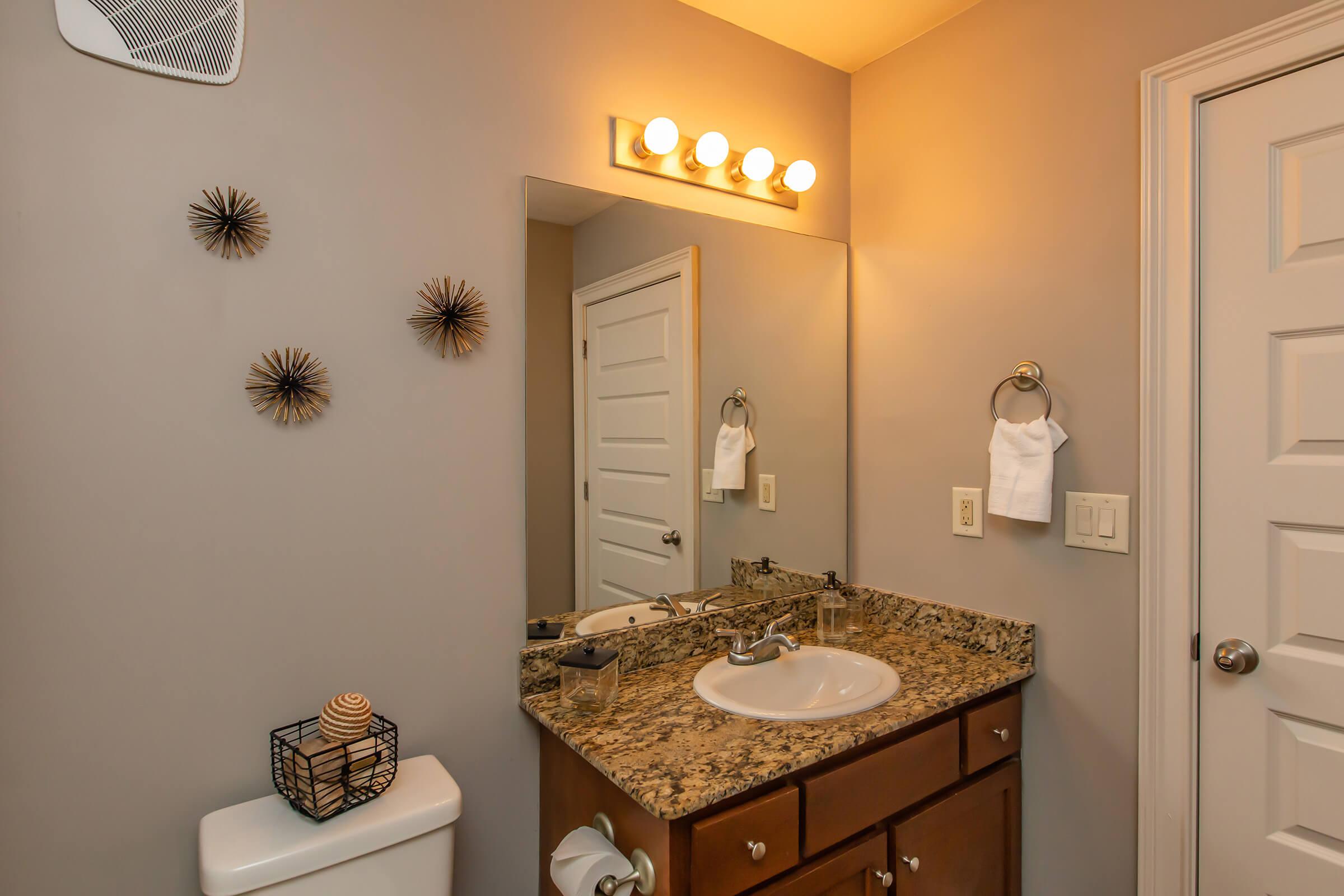 A modern bathroom featuring a granite countertop with a sink, a wall-mounted mirror, and decorative items. There is a toilet with a decorative container nearby, and two hand towels hanging on a towel rack. The walls are painted in a soft color, and there is stylish lighting above the mirror.