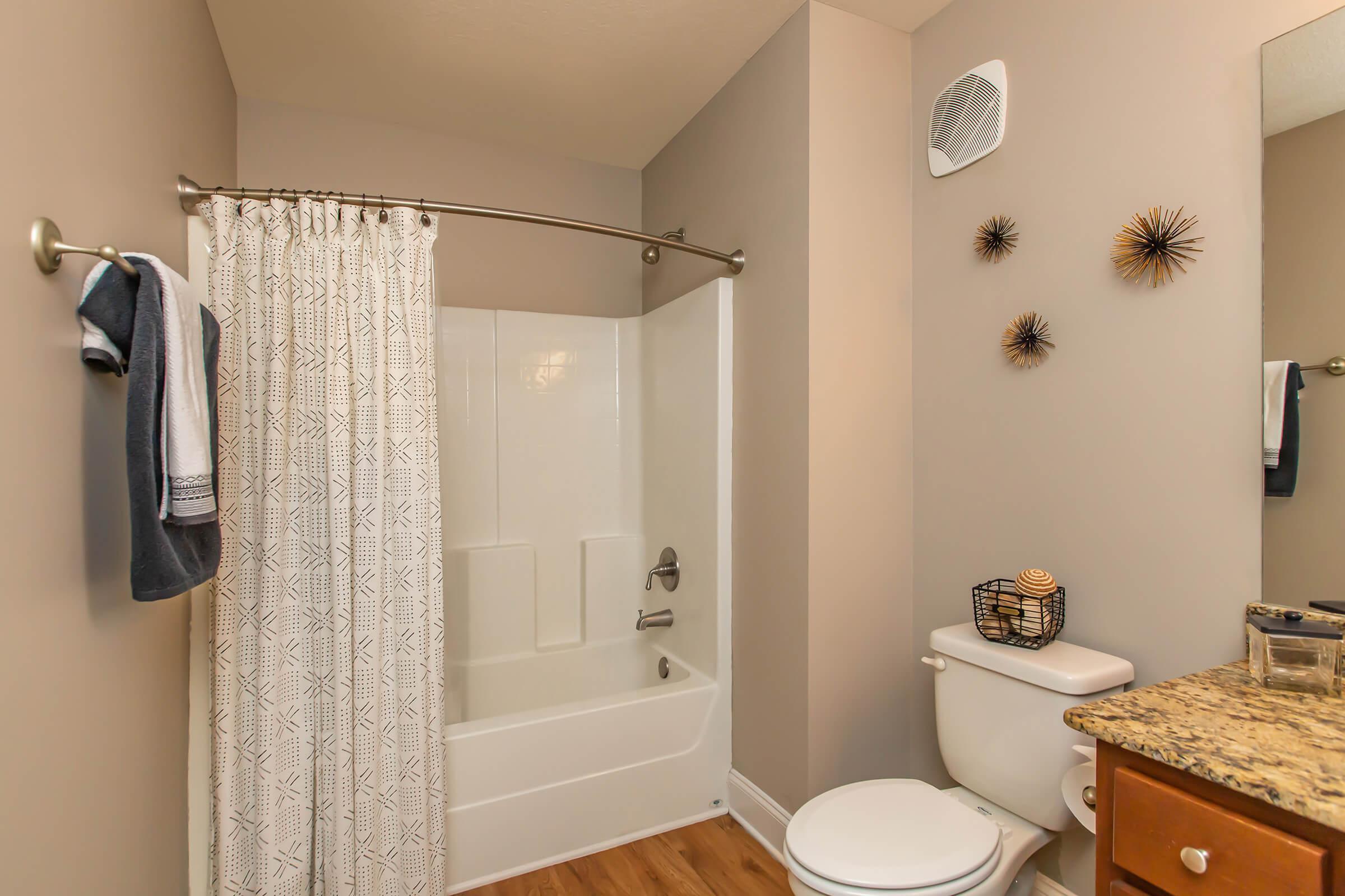 A modern bathroom featuring a shower/bathtub combination with a white curtain, a toilet, and a vanity with a granite countertop. The walls are painted light gray, and there are decorative wall accents, including a small basket and stylish wall ornaments. A towel hangs on a rail.